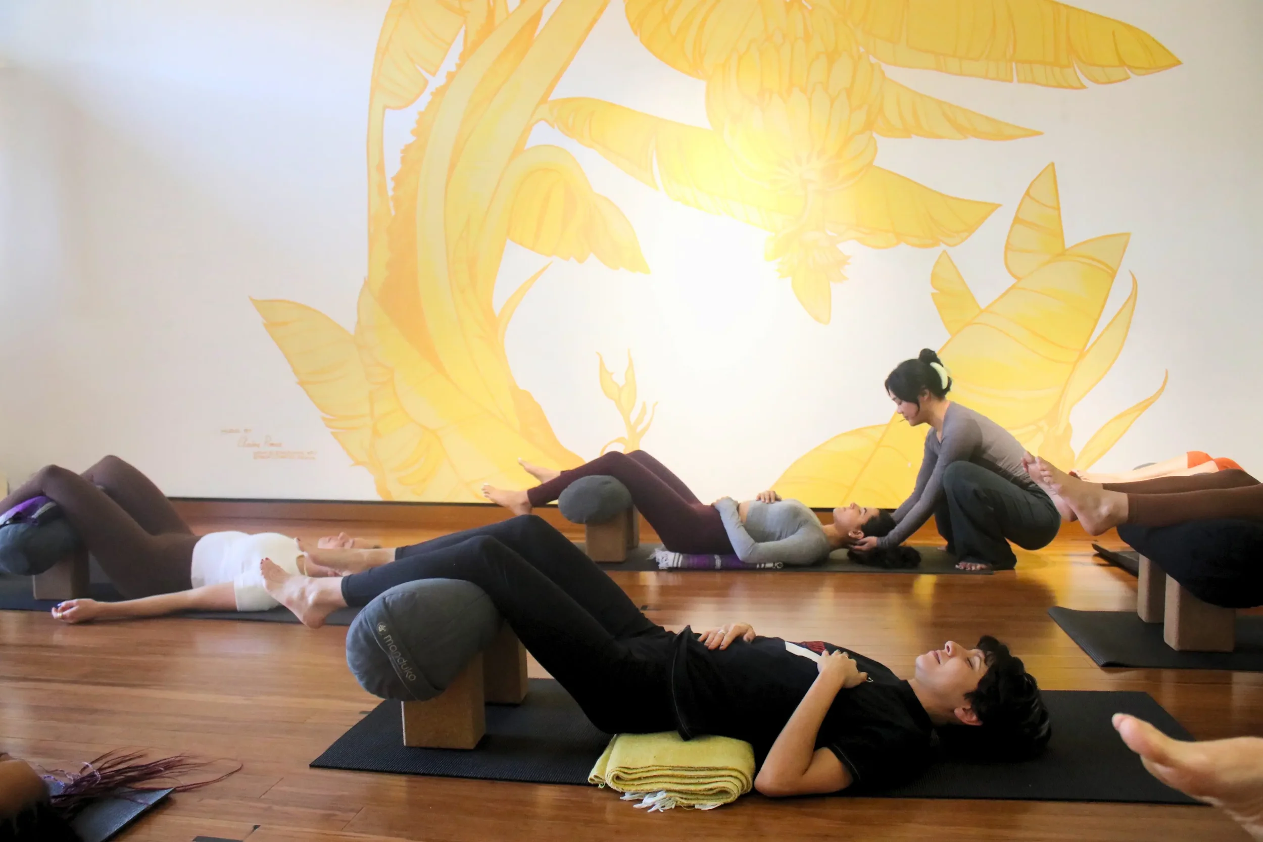 Group yoga class practicing a restorative posture lying on mats with legs elevated on bolsters supported by blocks. An instructor gently adjusts a student's head near a large yellow banana leaf mural.