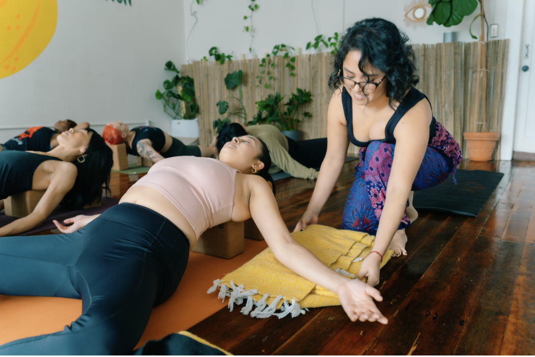 Group of female students in a restorative yoga/yin yoga class. The are lying on their backs in butterfly pose, supported by bolsters, blocks, and mexican blankets. The instructor is offering a supportive adjustment to a student's props.
