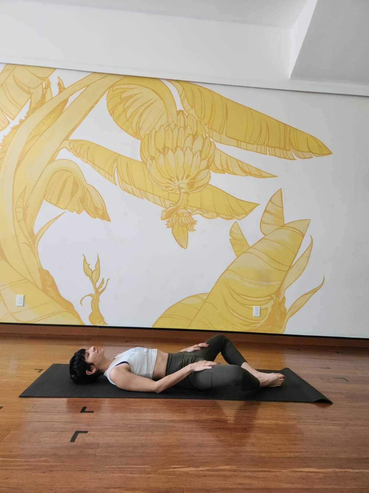 woman with a short pixie haircut wearing a white cropped tank top and fitted olive green leggins lies on her back with her feet together and knees open in a butterfly pose