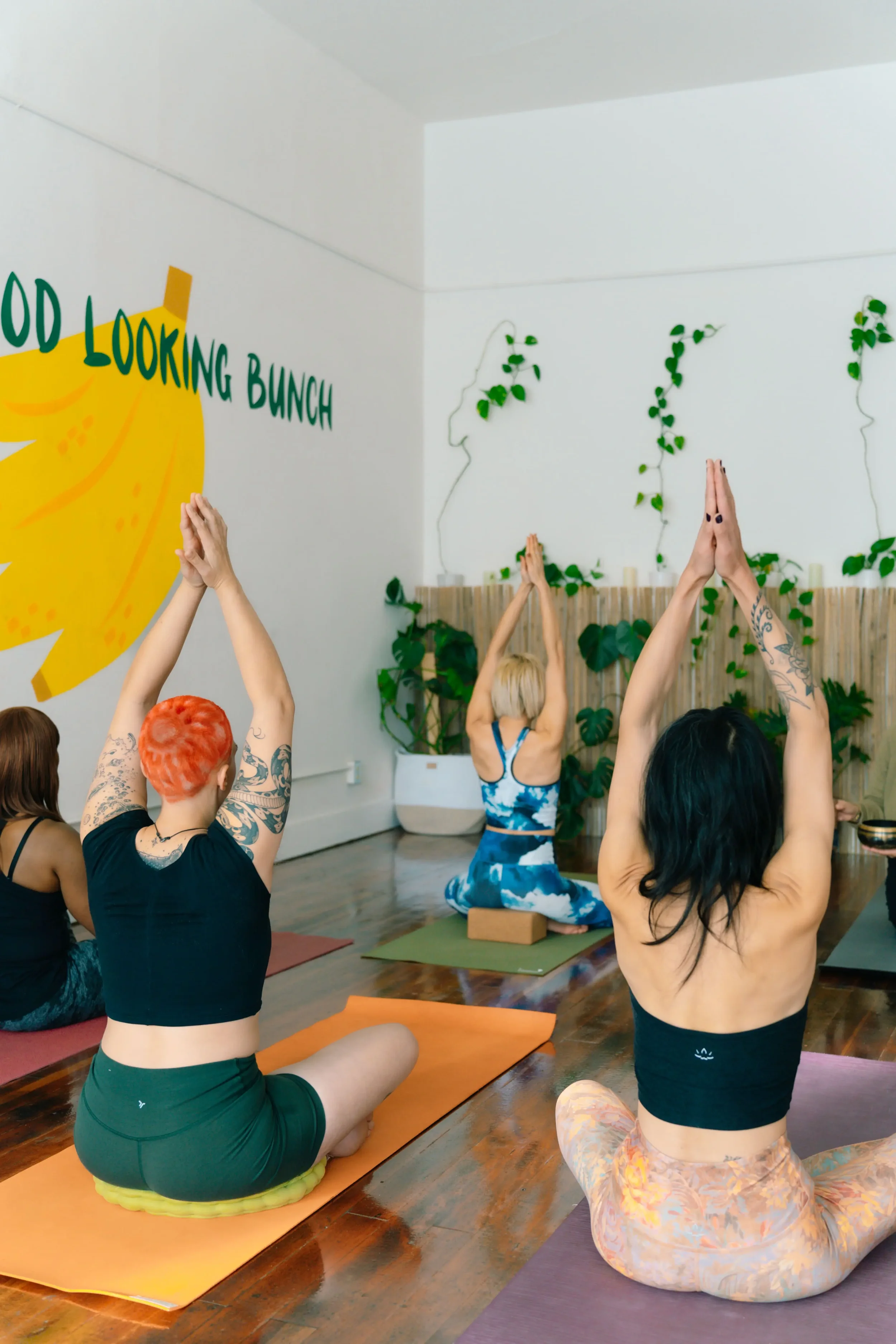 Four women in a bright yoga studio. They sit cross-legged on colorful mats with their hands pressed together above their heads in prayer position.