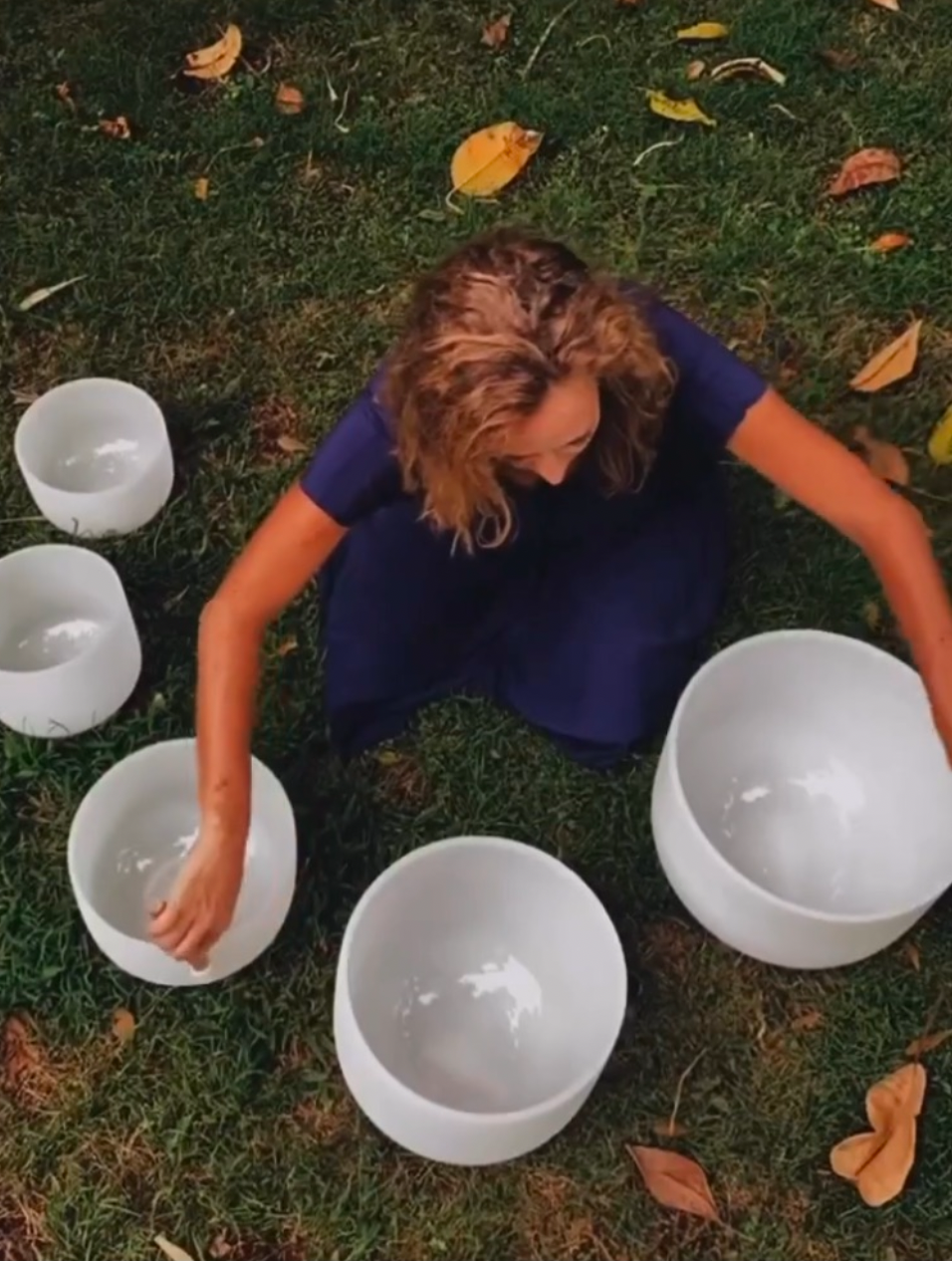 A woman sitting on grass surrounded by white crystal singing bowls, with a few fallen yellow and orange leaves scattered around.