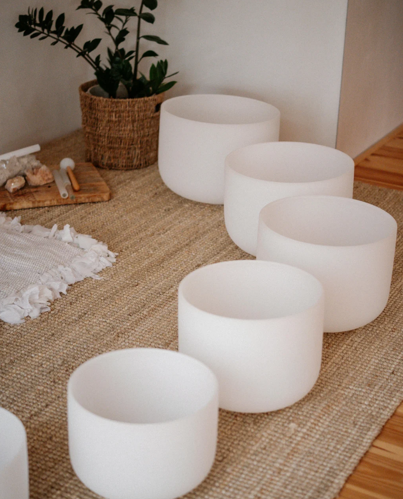 Five white singing bowls arranged in a line on a woven rug, with a potted plant and a wooden board with rocks and a tuning fork nearby.