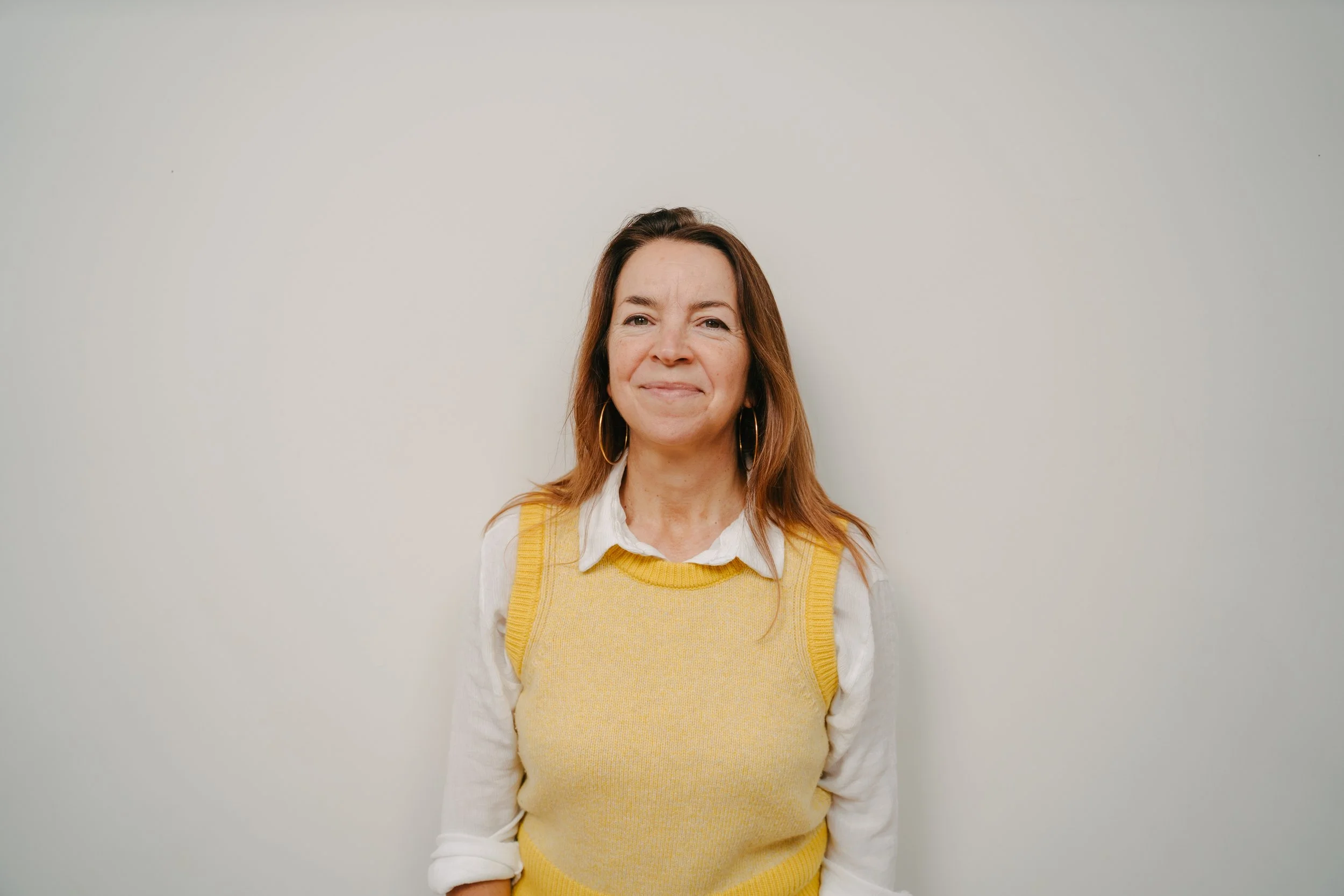 Women with brown hair wearing a white shirt and yellow vest, standing against a light-colored wall. Credit Emilie Lashmar.