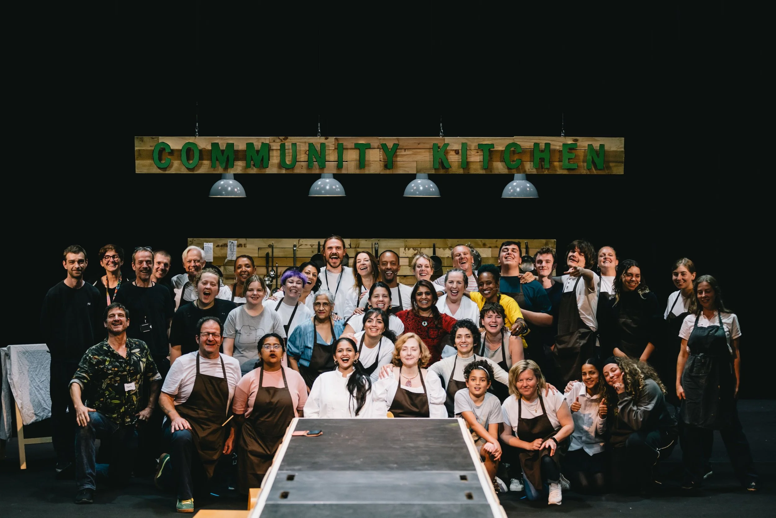 Group of diverse people smiling and posing in front of a sign that says 'Community Kitchen' with cooking utensils hanging behind them. Image courtesy of Brighton People's Theatre of Brighton Festival production of Born and Bread. Credit Rosie Powell.