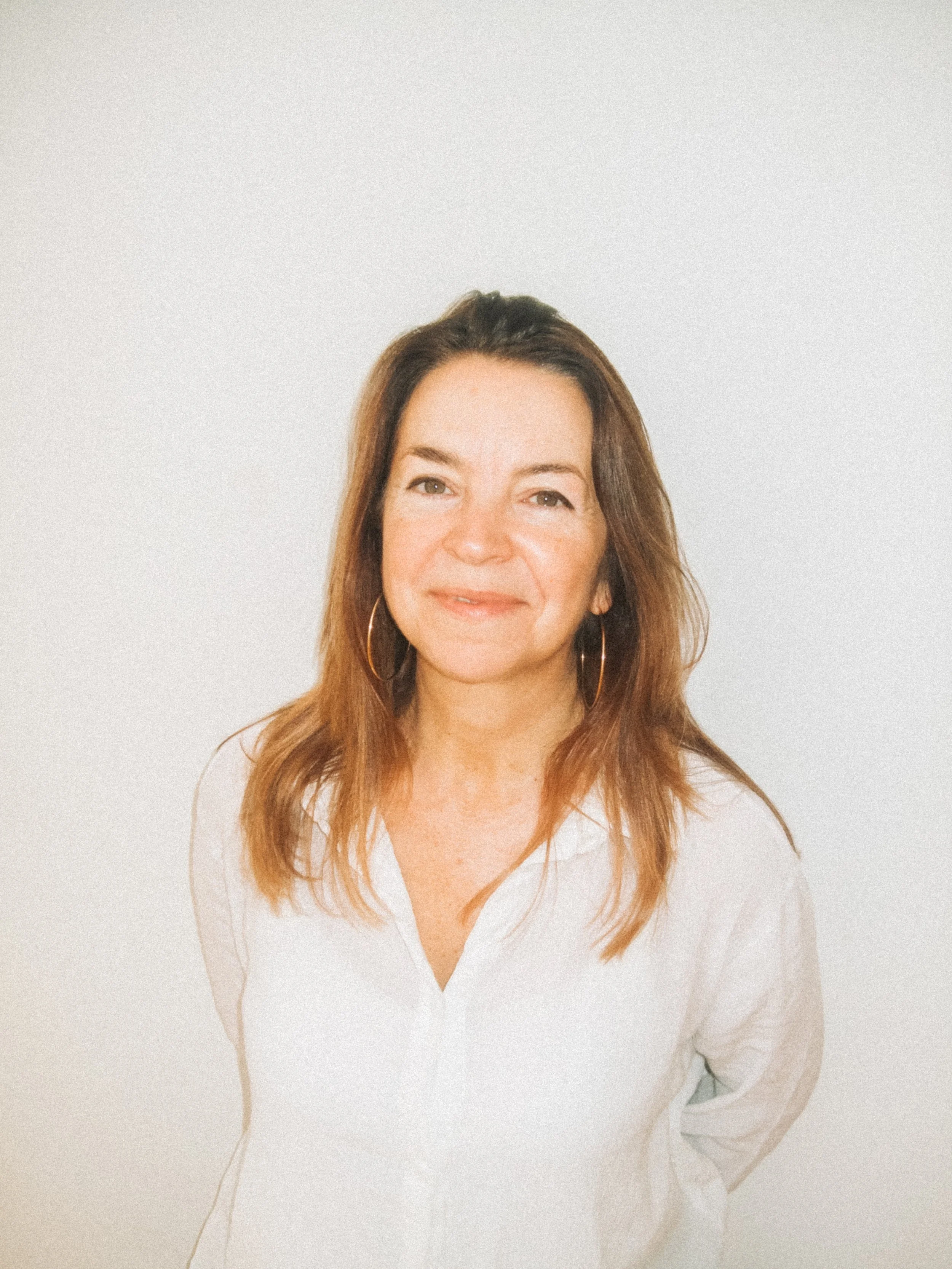 A woman with brown hair and hoop earrings, wearing a white button-up shirt, smiling while standing against a plain white background. Credit Emilie Lashmar.