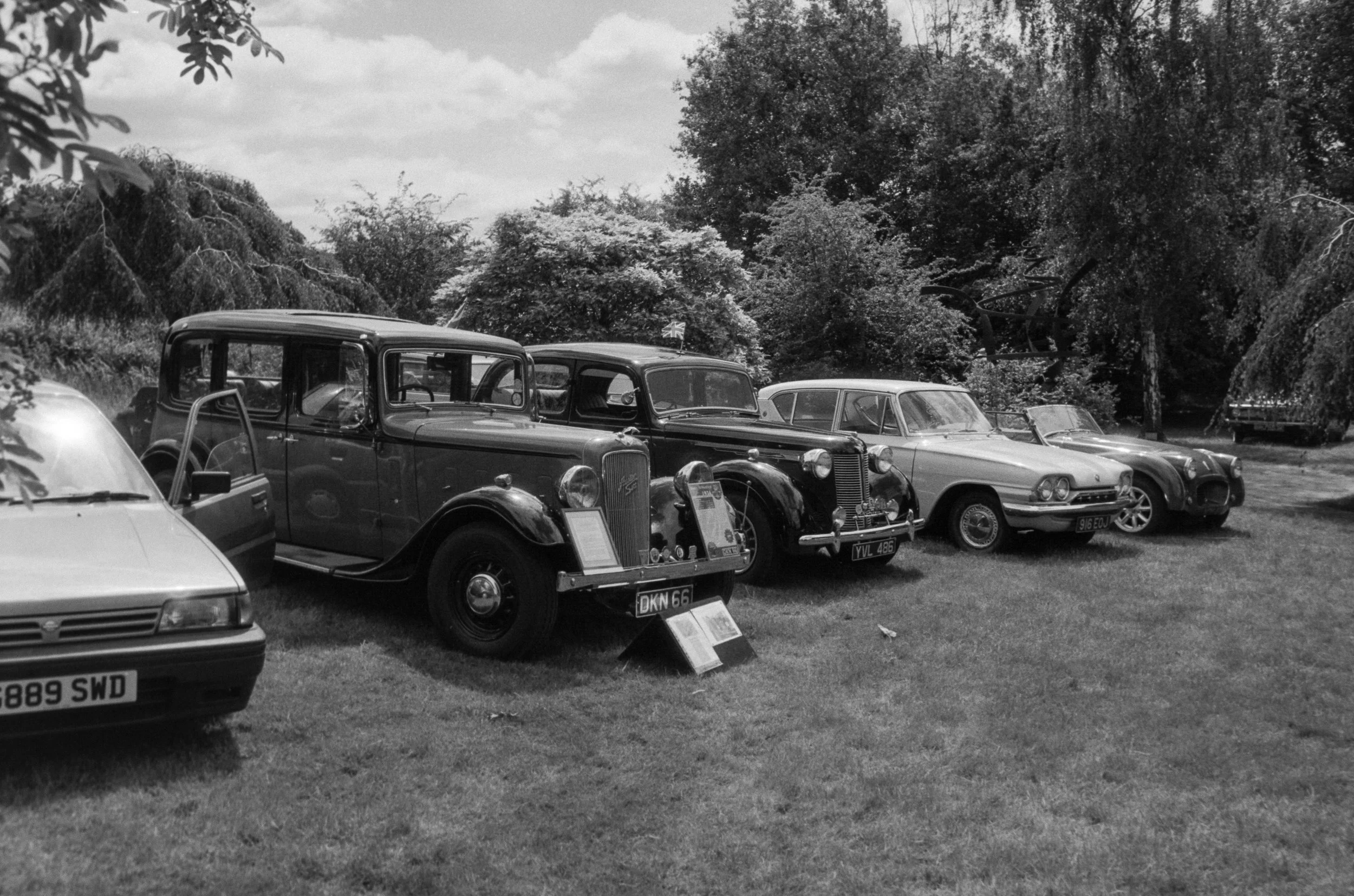 A black and white photograph of a vintage car exhibition outdoors, with multiple cars parked on a grassy area under trees, and informational plaques in front of some cars.
