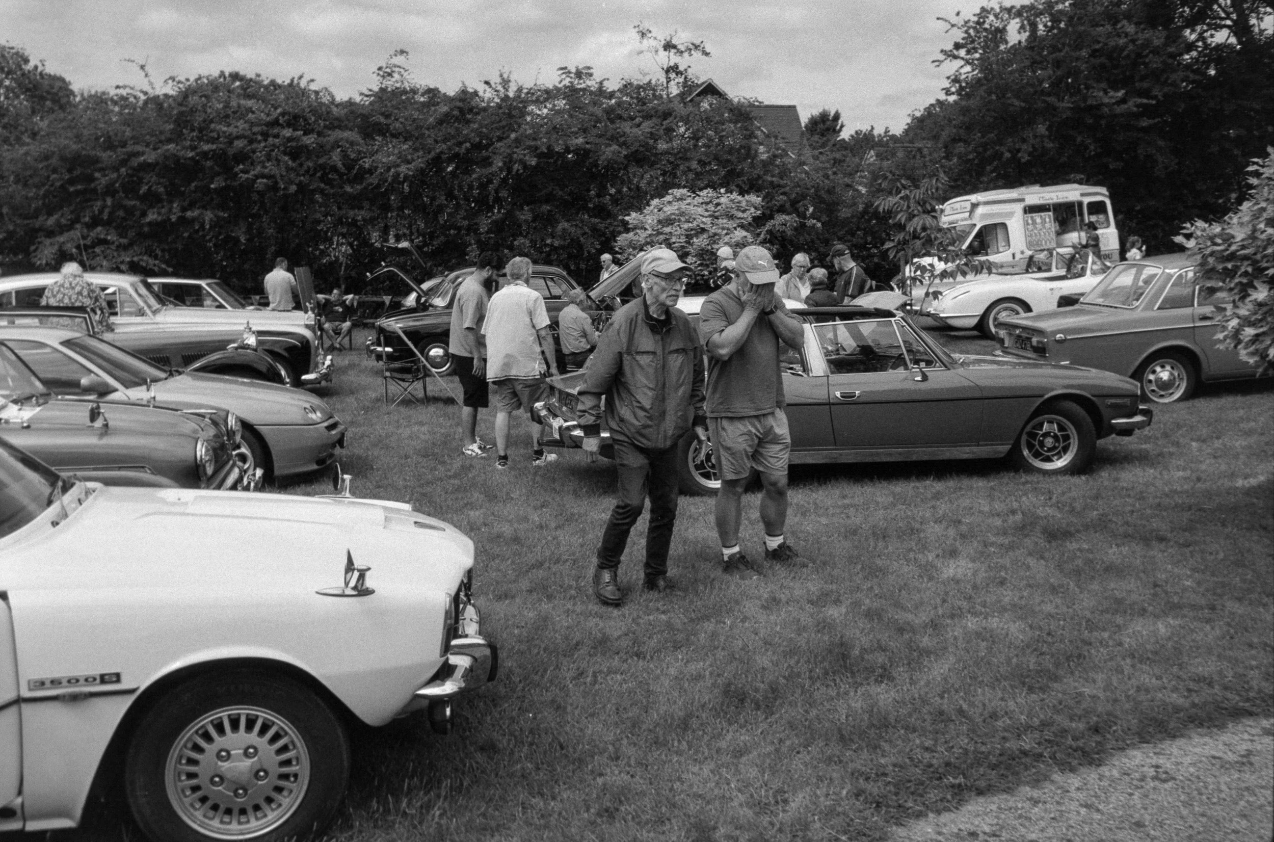 Black and white photo of a classic car show in a grassy park with people examining vintage cars, some with hoods open, and trees in the background.