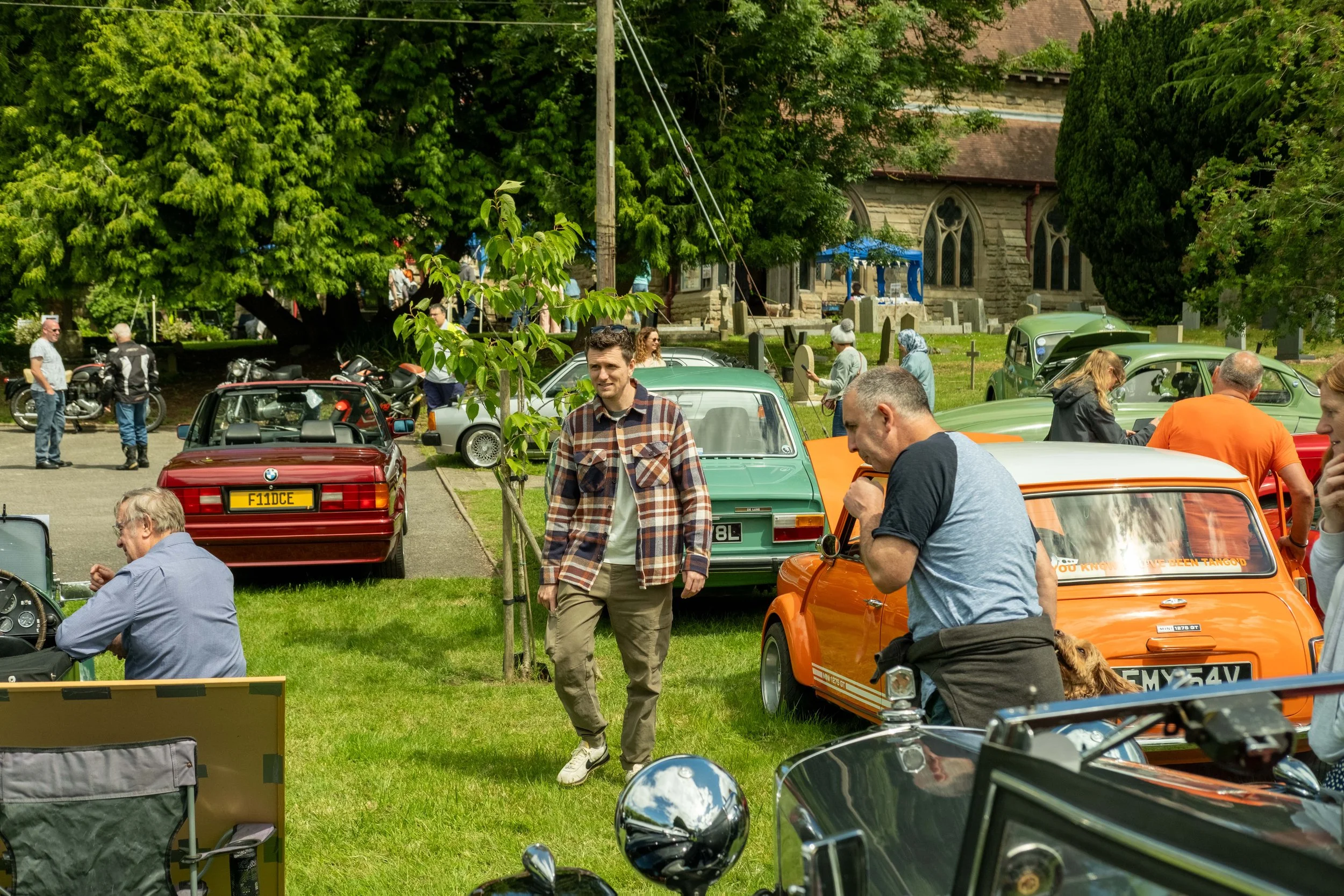 A car show featuring vintage and classic cars with people walking around and viewing the vehicles in a park-like setting with large trees and a stone building in the background.