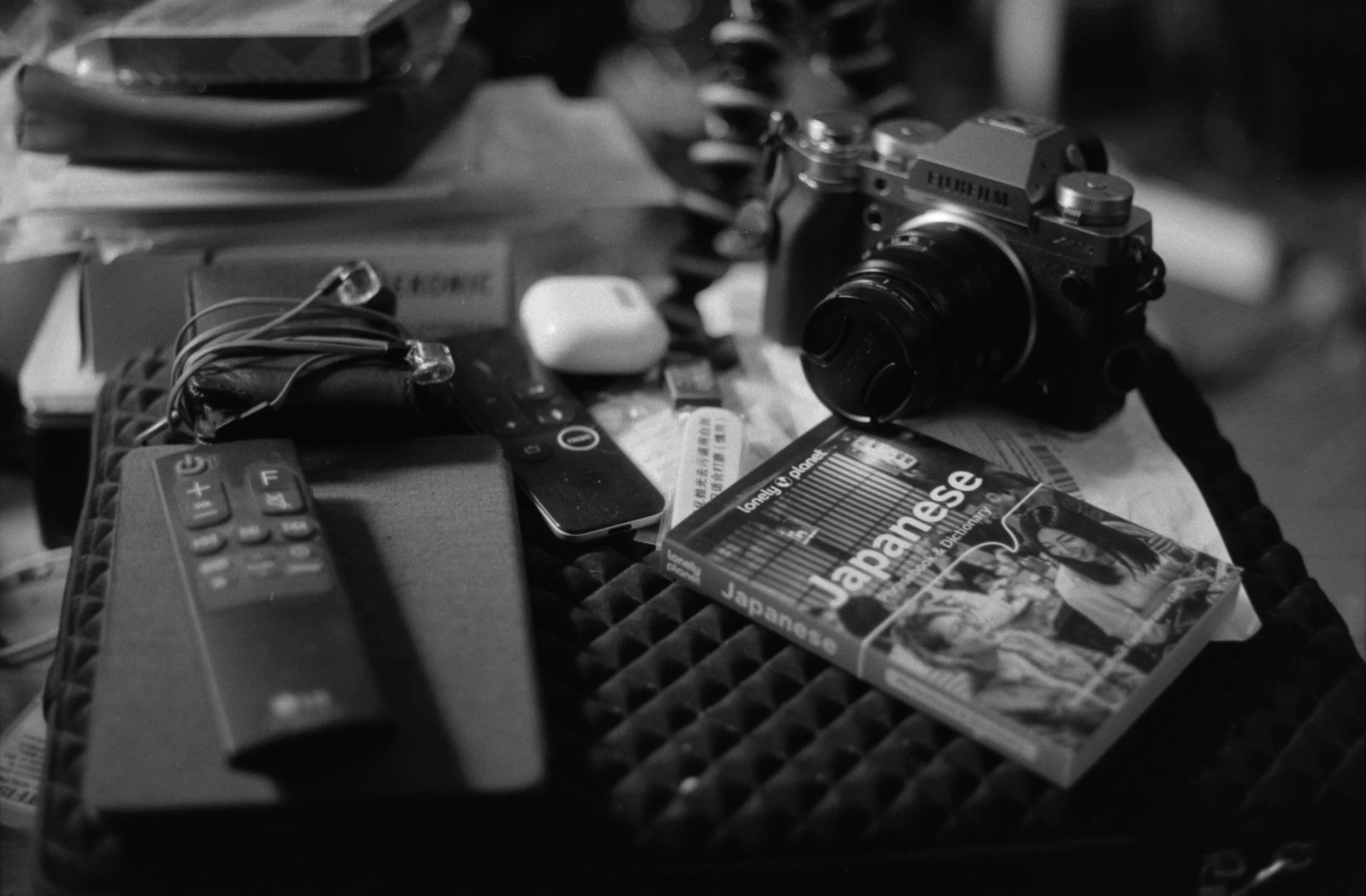 Black and white photo of a cluttered table with a digital camera, remote controls, a magazine, earphones, and various objects.