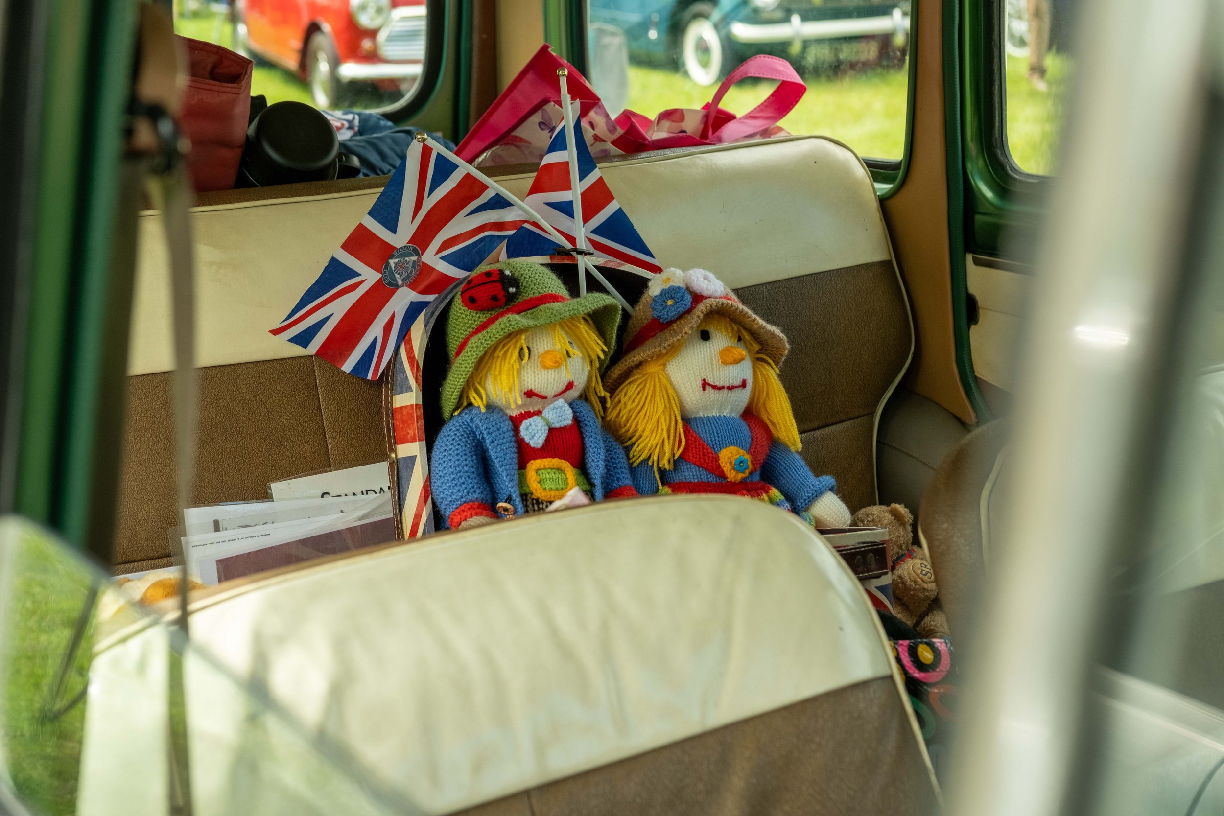 A display of handmade knitted dolls, one with a ladybug hat, next to small British flags and various bags inside a vehicle, with vintage cars visible through the windows.