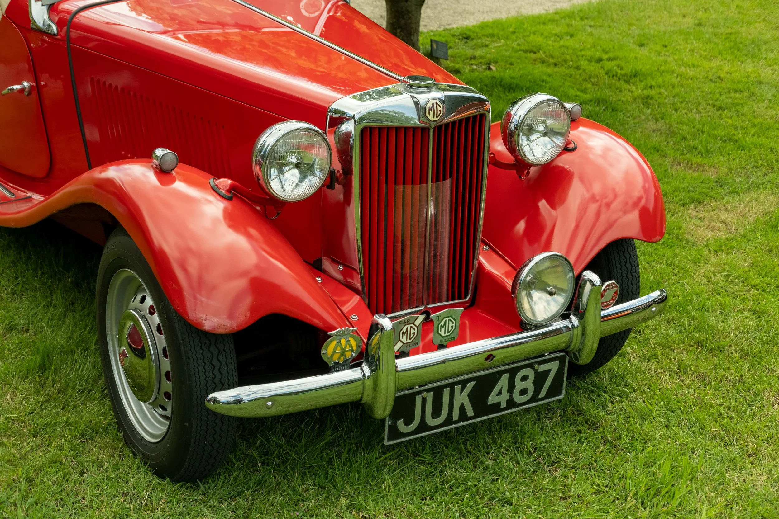 A close-up of a vintage red MG car parked on grass, showing front grille, headlights, bumper, and license plate.