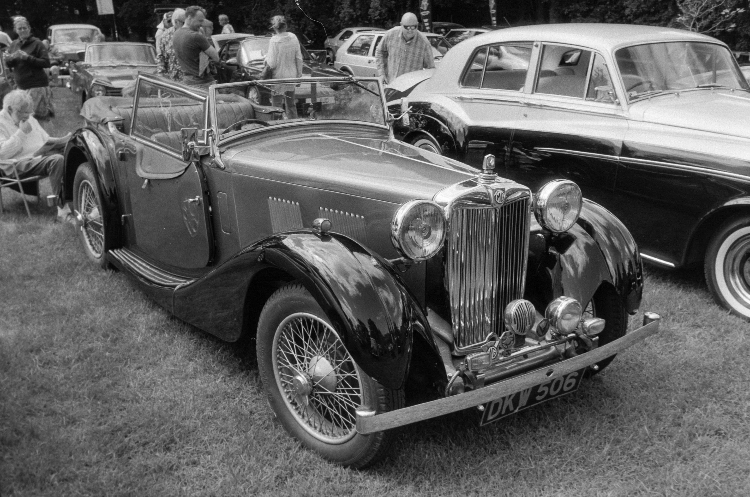A vintage car show with a classic convertible MG car in the foreground and other vintage cars in the background, surrounded by people viewing and chatting at an outdoor event.
