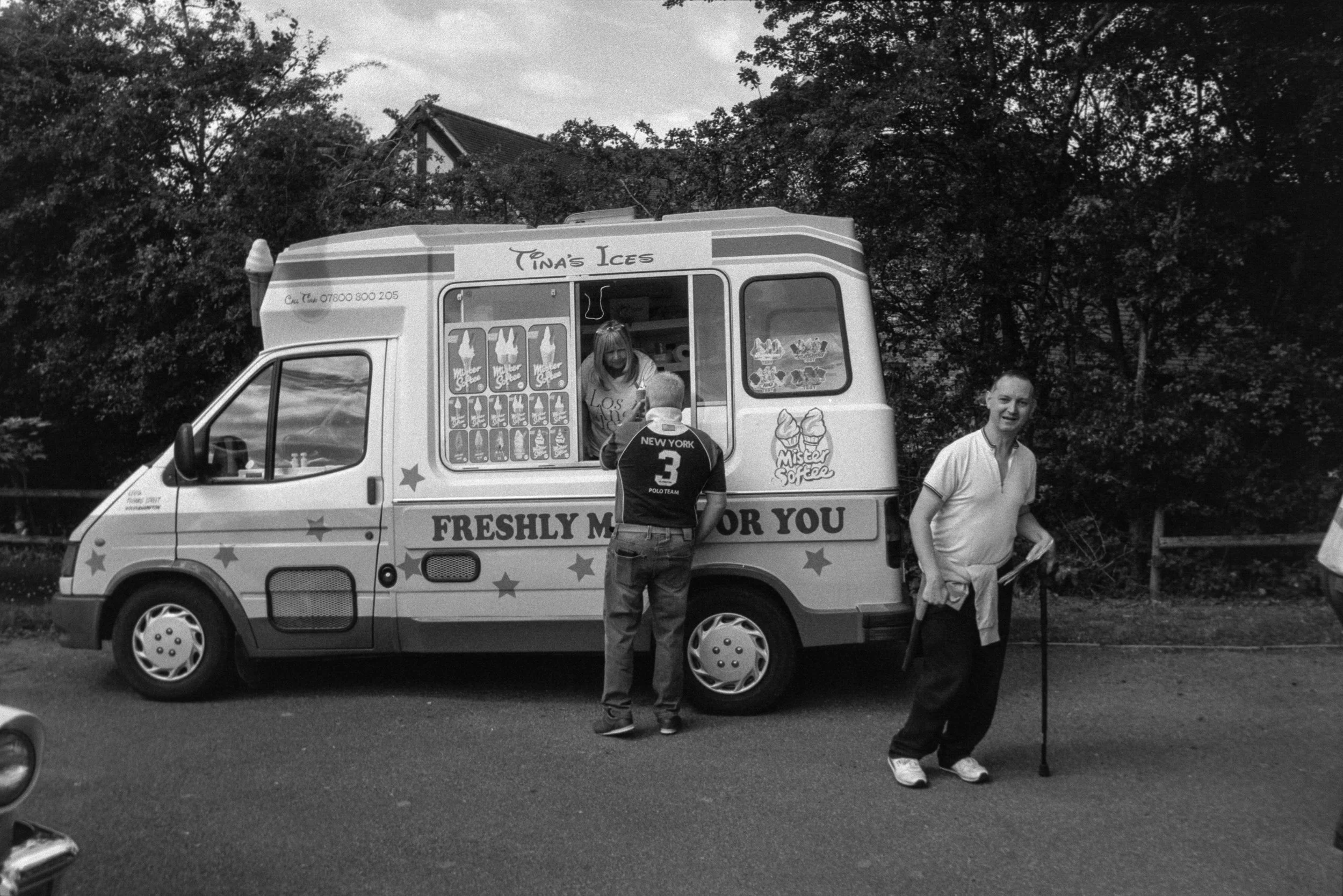 A black and white photo of a small ice cream truck with the name "Tina's Ices" on the top. There are two men standing in front of the truck, one wearing a sports jersey and the other holding a cane. The truck has a sign that says "Freshly Made" and d