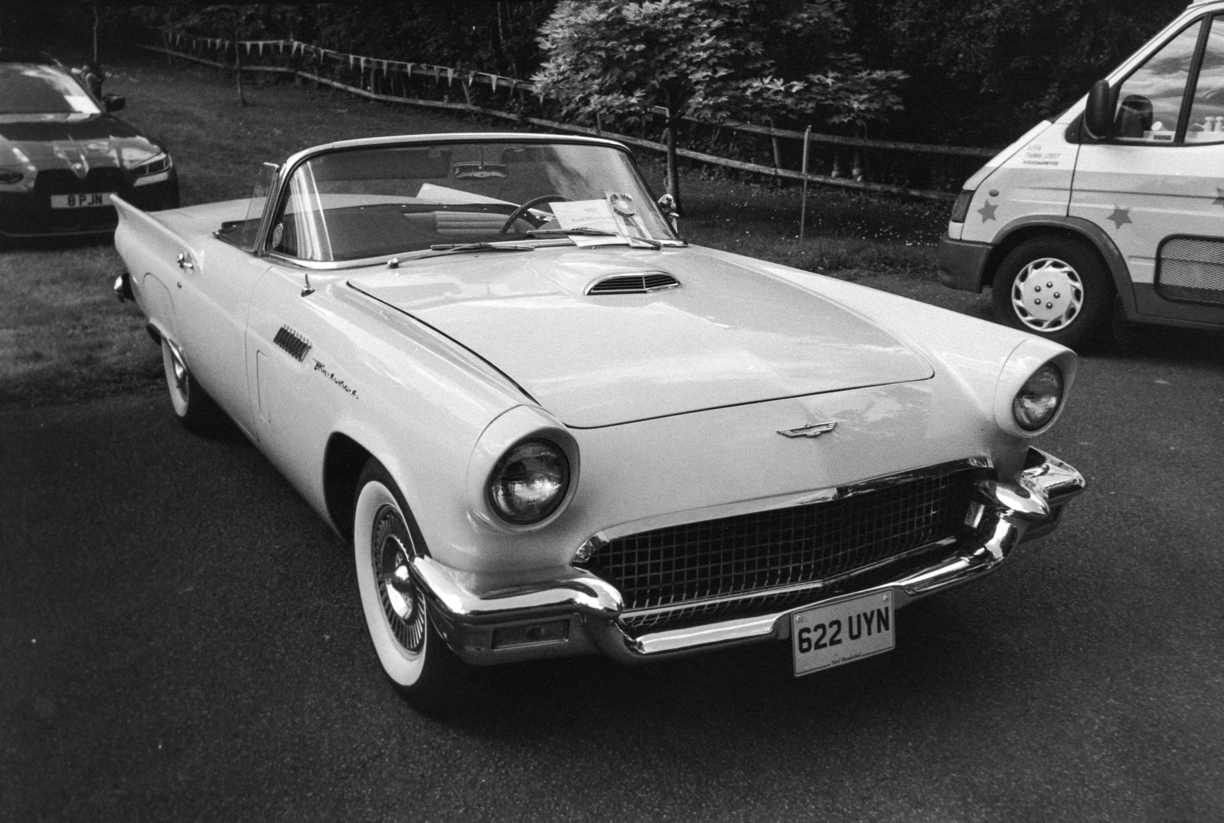 A vintage white Chevrolet Corvette convertible car parked on grass, with other vehicles and trees in the background.
