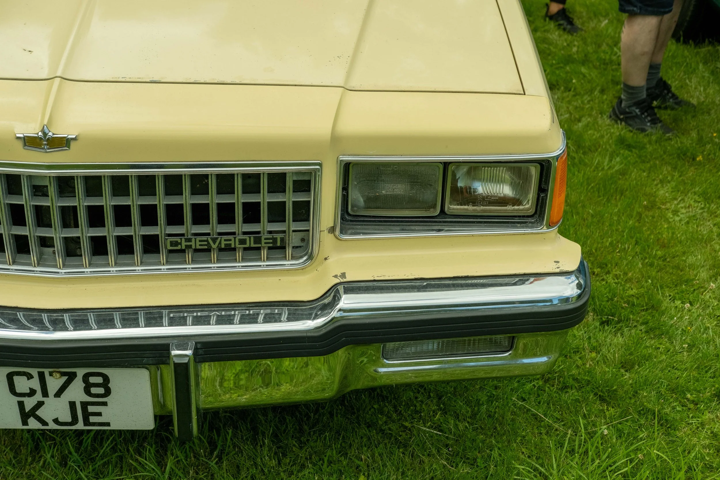Close-up of the front of a vintage yellow Chevrolet car with a Chevrolet emblem on the grille, set on grass with a person's legs in the background.
