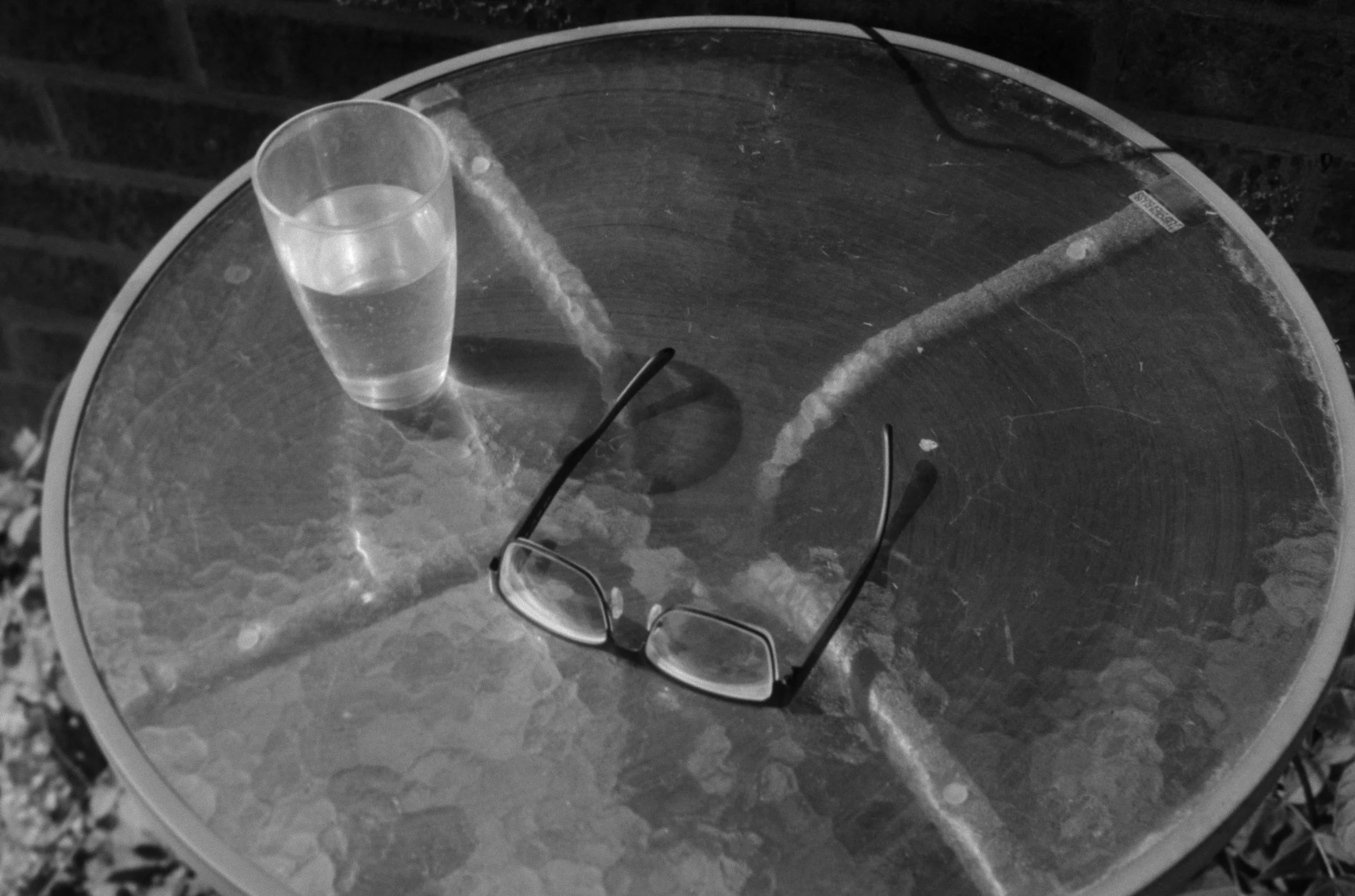 A clear glass of water and a pair of eyeglasses resting on a round glass outdoor table with a textured surface.