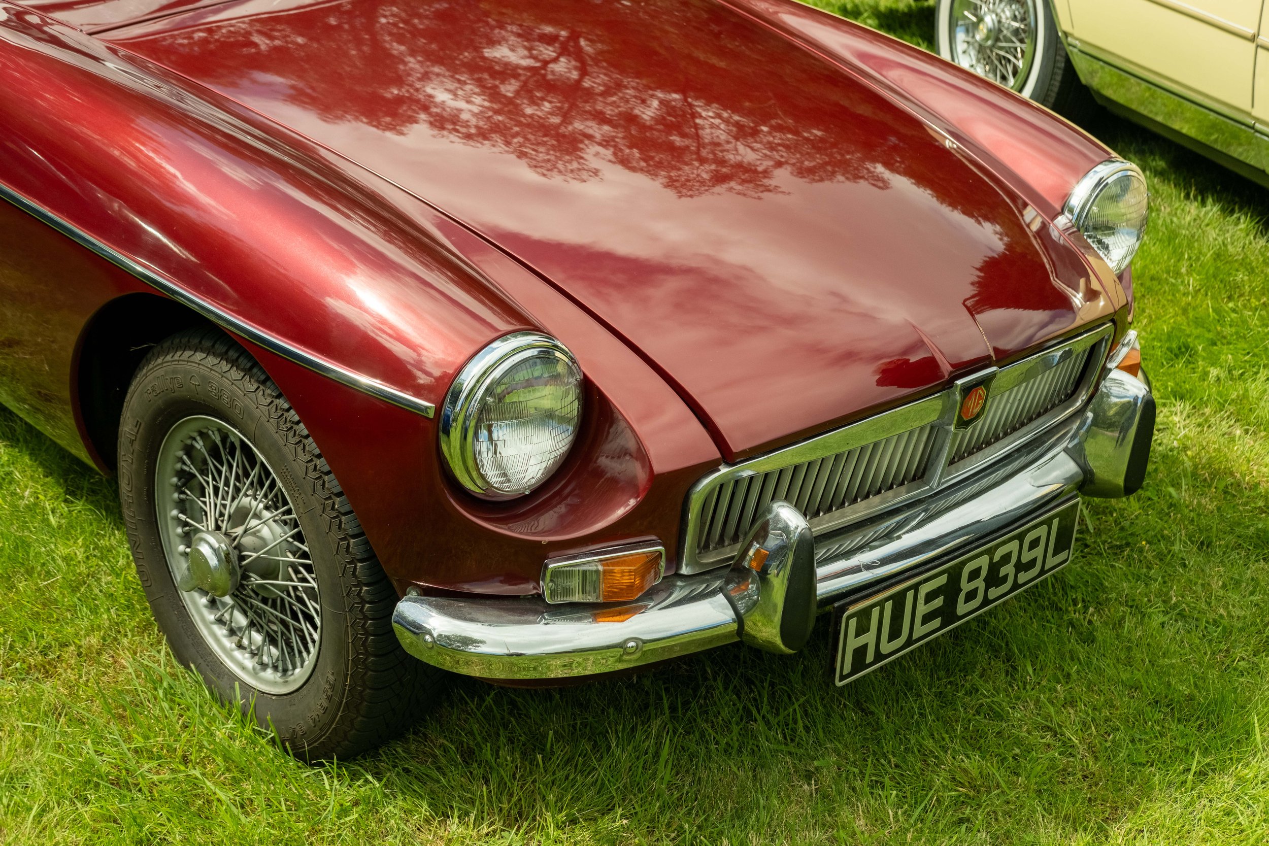 A classic red vintage car parked on the grass, with part of another vintage car visible nearby. The car has a shiny exterior, wire-spoked wheels, and a license plate reading 'HUE 839L'.