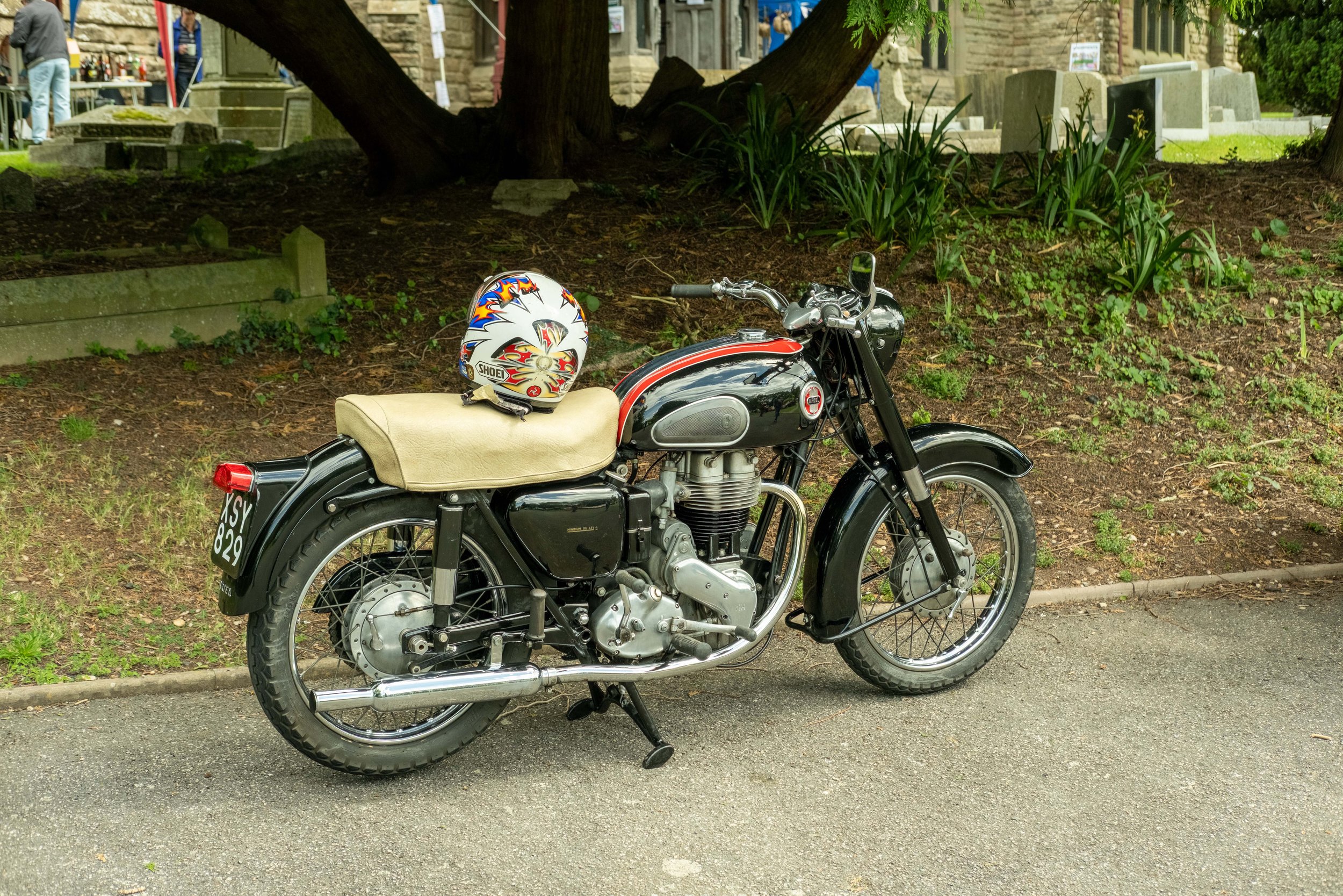 A vintage black motorcycle with a beige seat and a colorful helmet resting on the seat, parked on a street near a grassy area with trees and plants. In the background, there are people, buildings, and outdoor market stalls.