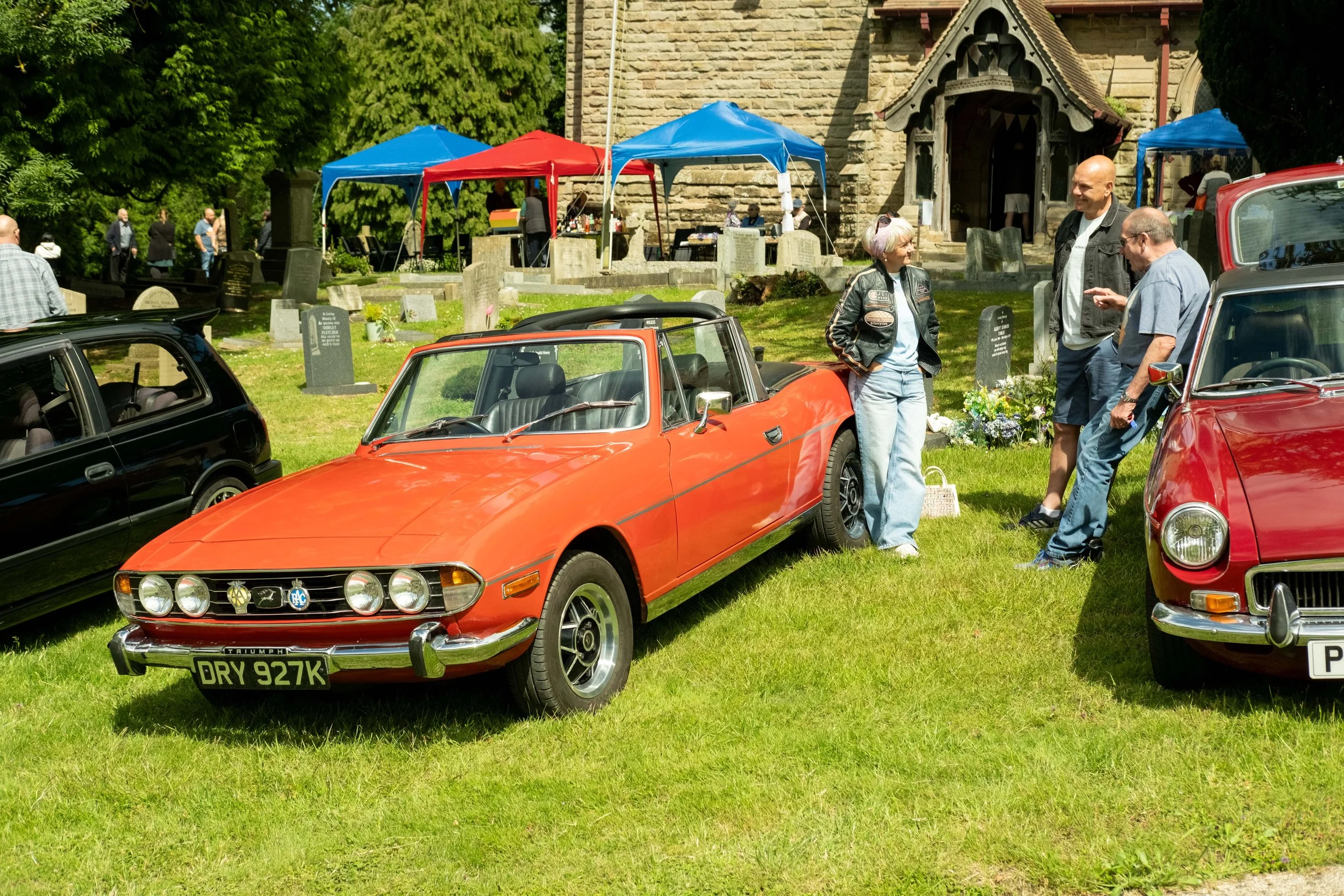 People at a car show in a grassy area outside a church, with vintage cars and tents in the background.