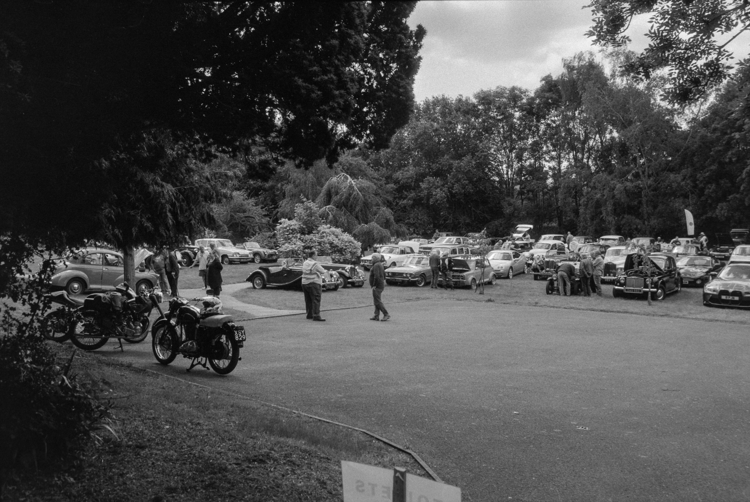 Black and white photo of a vintage car show in a park with people walking around and trees in the background.