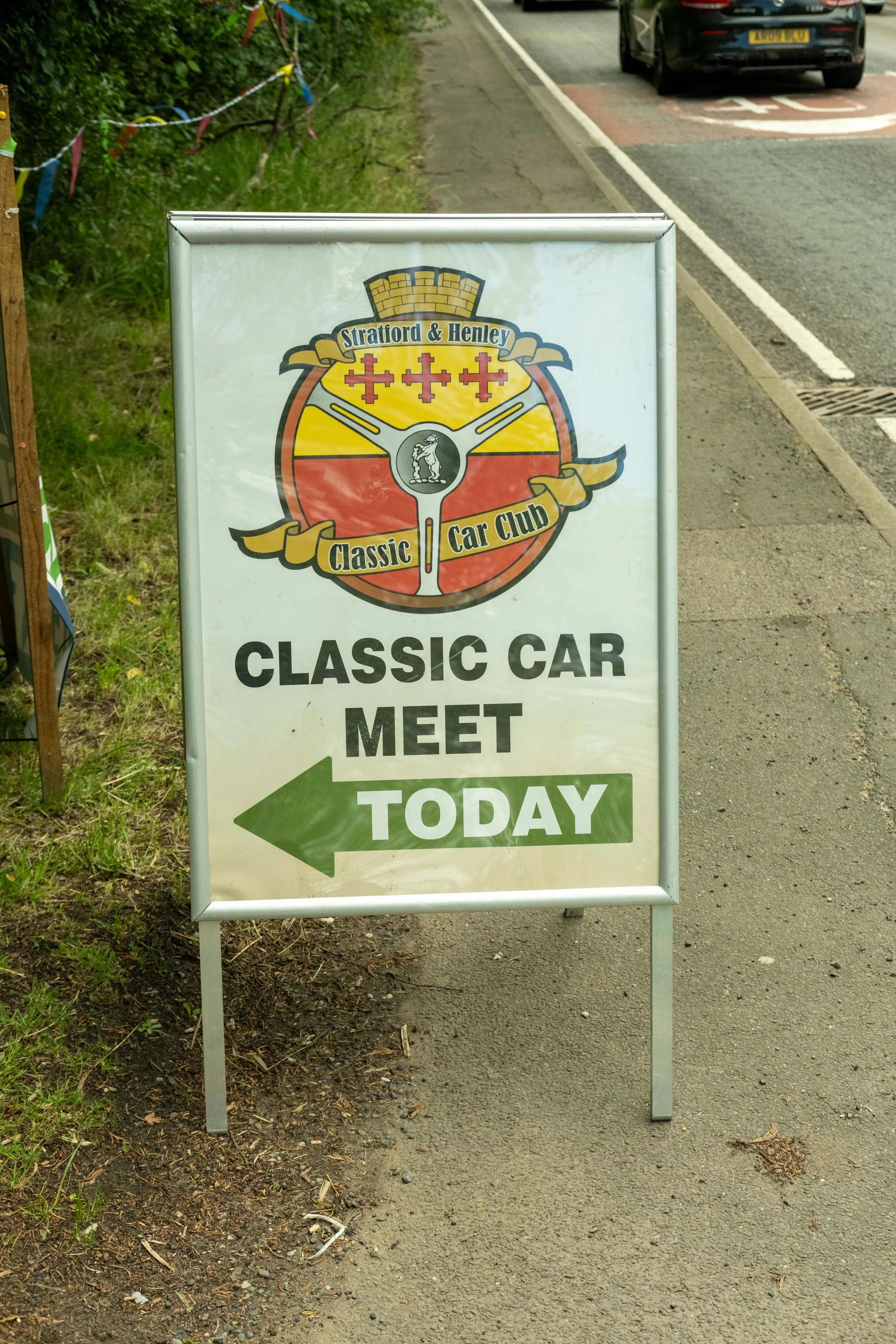 A sidewalk sign for the Stratford & Henley Classic Car Club pointing to the left for a meet today, with a red, yellow, and black emblem featuring three red crosses at the top, a woman in the center, and a yellow banner with black text.