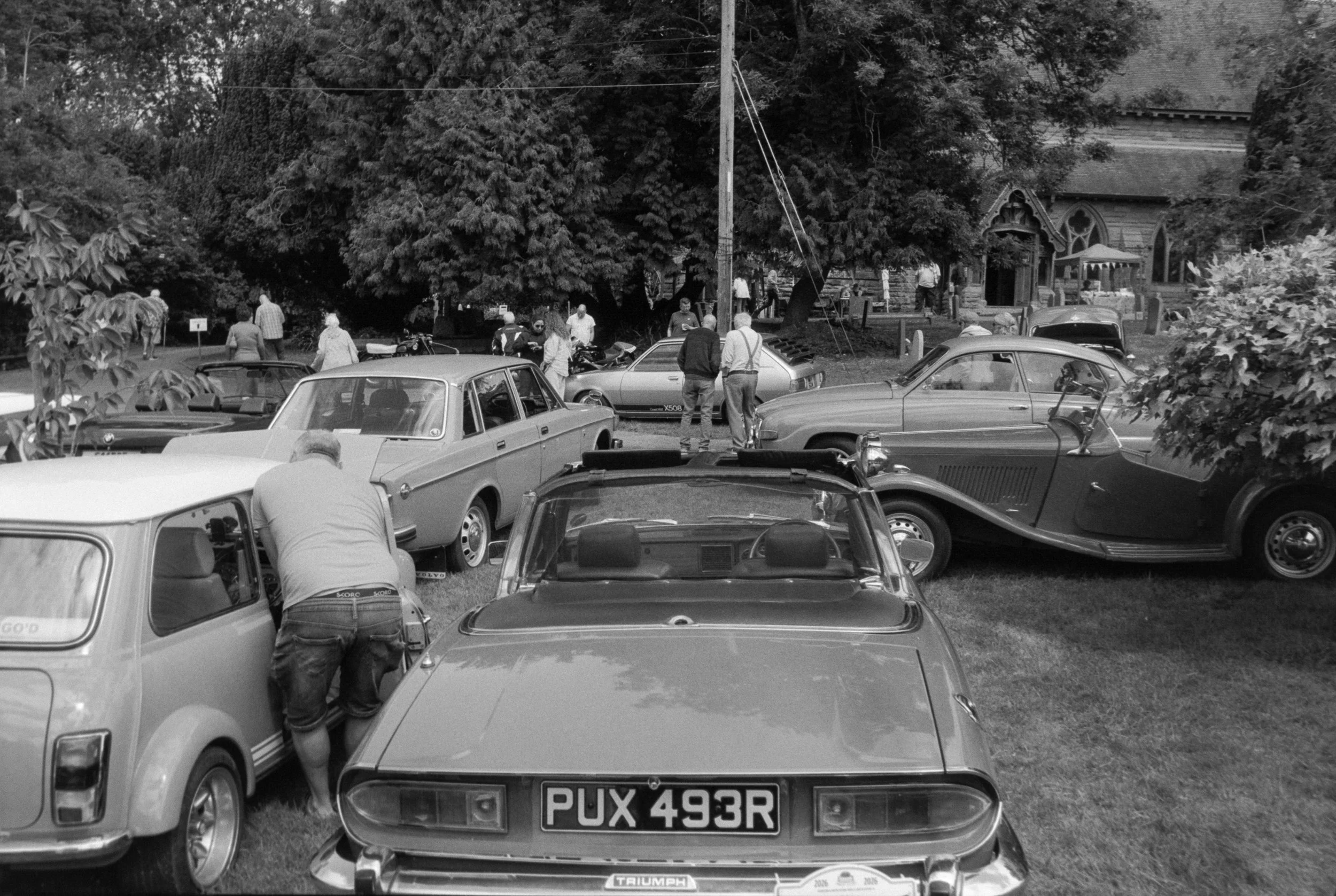 Black and white photo of a vintage car show with various classic cars parked on grass and people walking around, including some inspecting vehicles and talking, in a park-like setting with trees and a historic church in the background.
