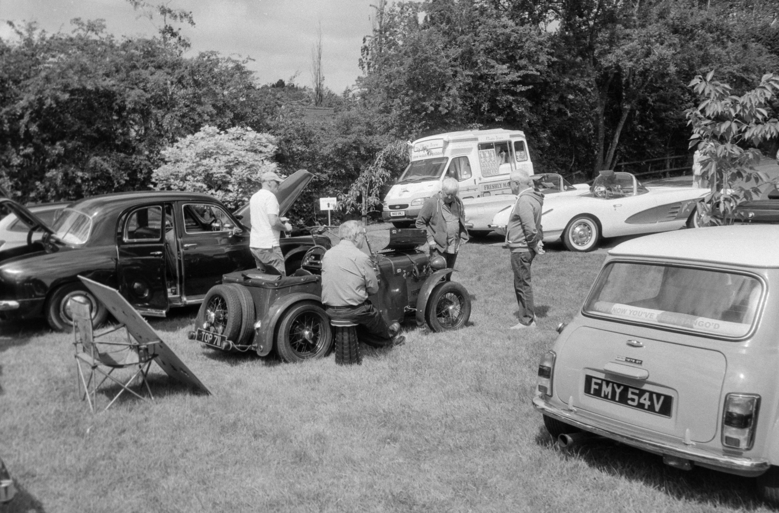 A black-and-white photo of a vintage car show with multiple classic cars parked on a grassy field and people inspecting the vehicles. Several trees are in the background.
