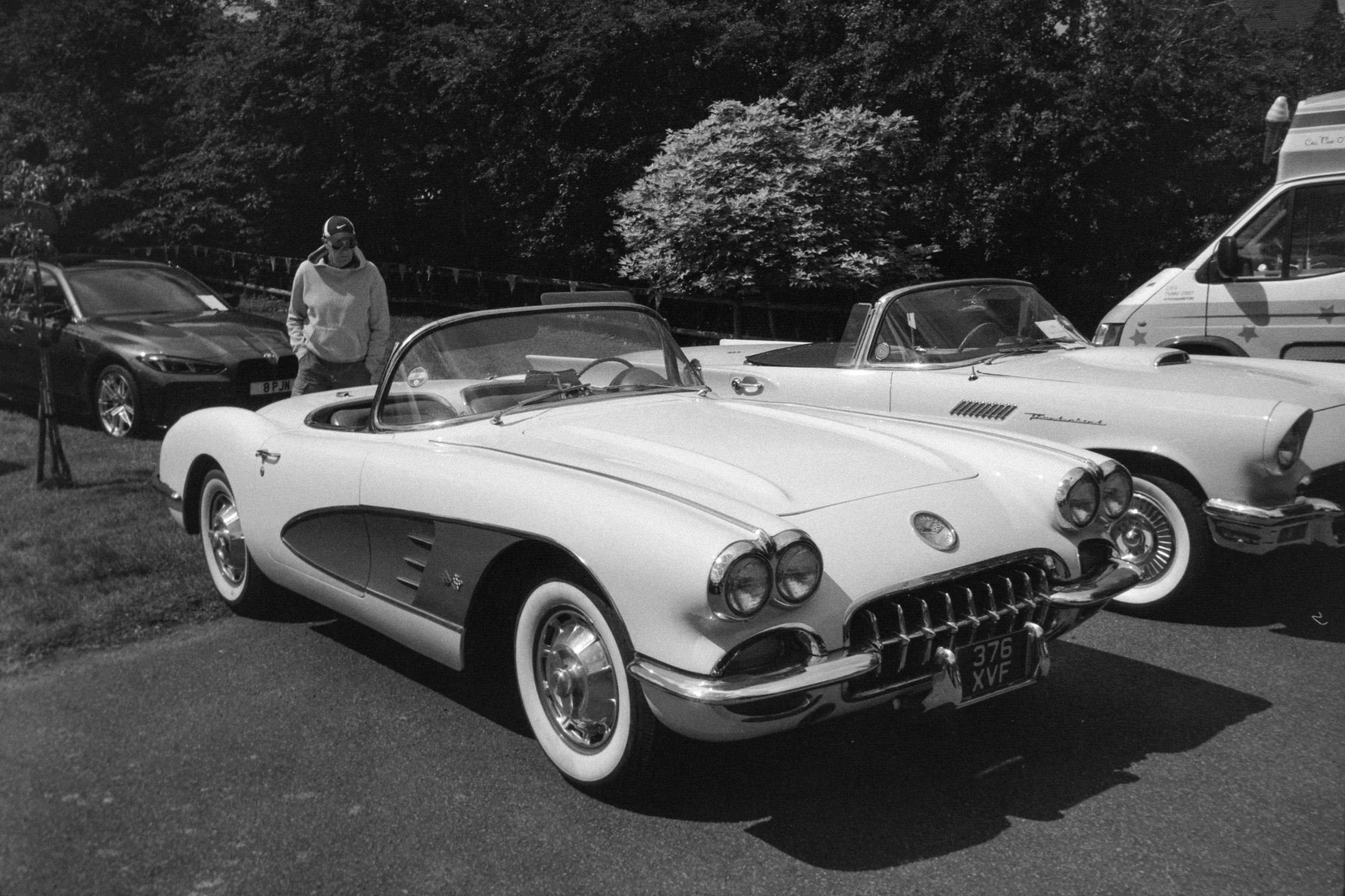 Black and white photo of vintage Chevrolet Corvette and modern cars parked outdoors, with a person standing near the classic car and trees in the background.