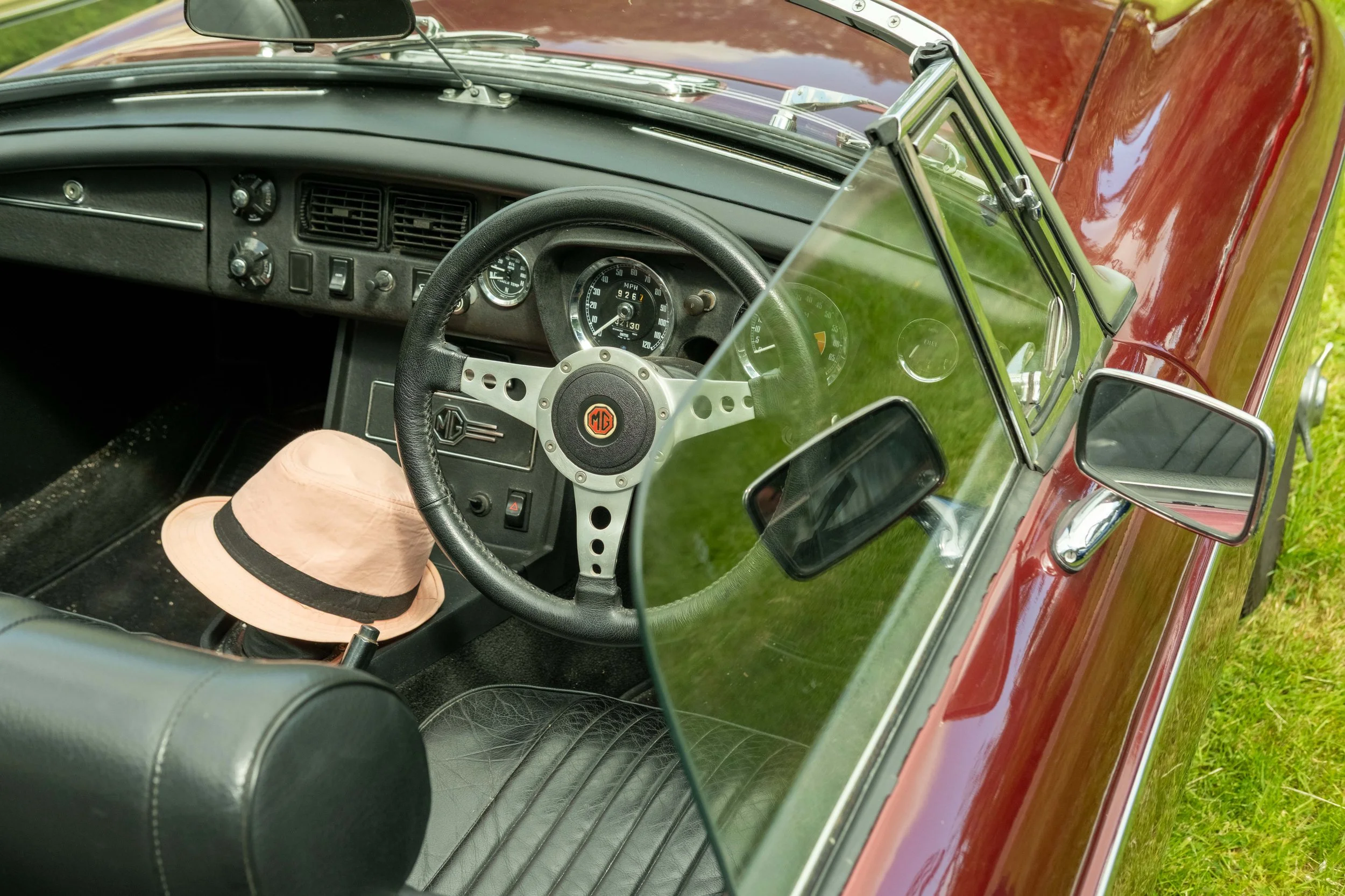View of a vintage red MG car's interior from the driver's side, showing the dashboard, steering wheel, and a light-colored hat on the seat.