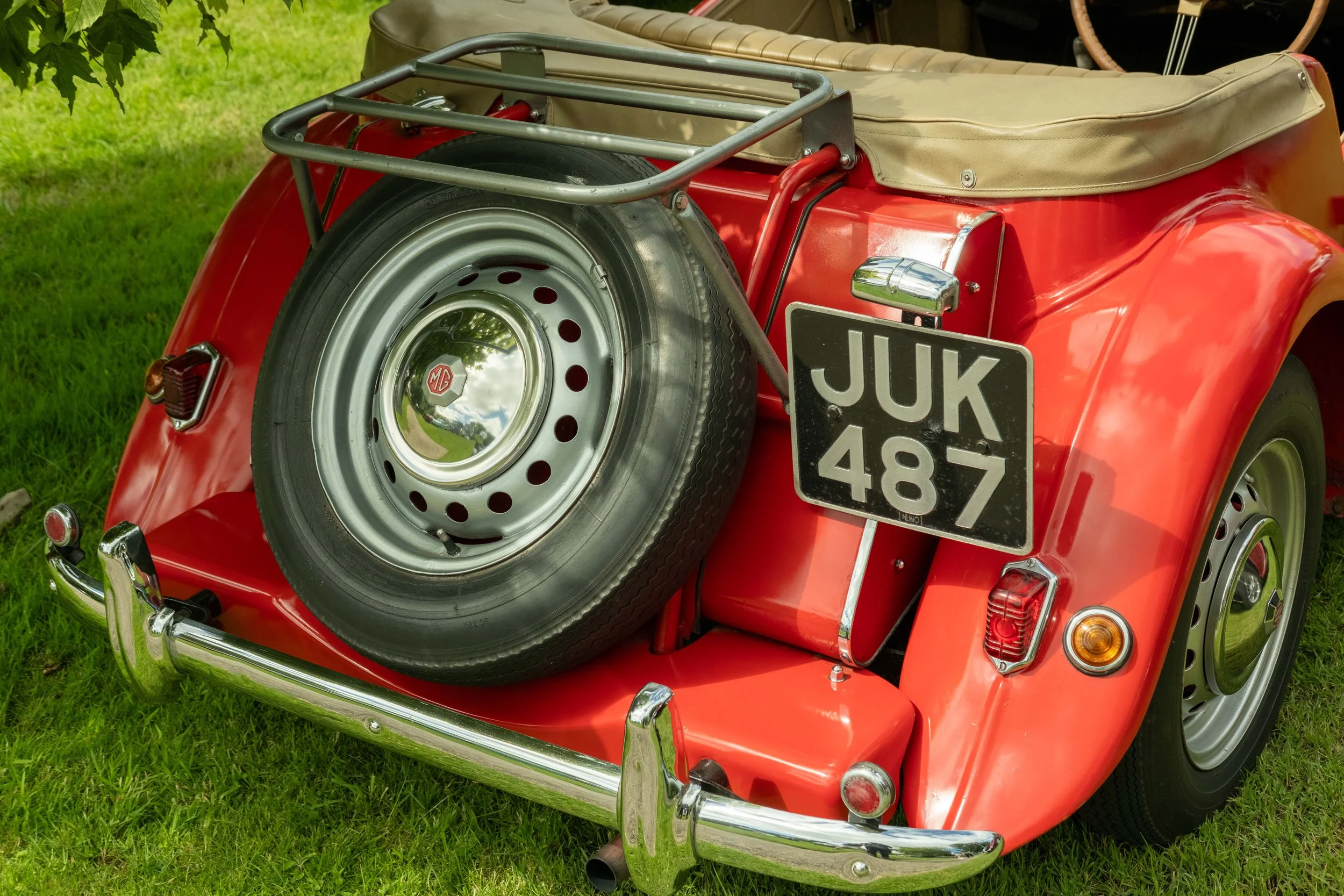 A vintage red convertible car with a spare tire mounted on the back and a metal luggage rack. The car is parked on green grass, and there is a beige soft top. License plate reads 'JUK 487.'