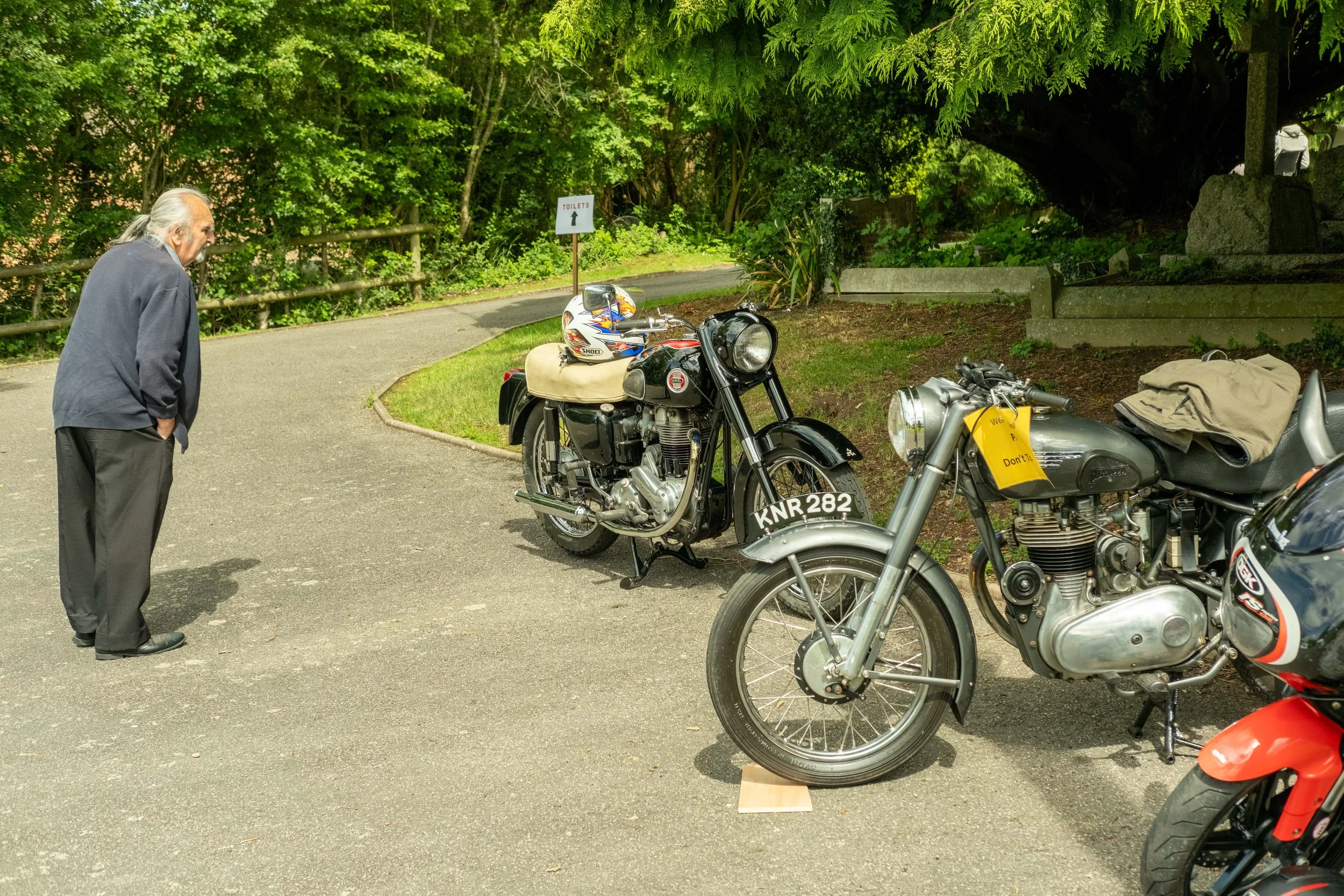 An elderly man with long gray hair tied back, wearing a dark gray jacket and black pants, looks at vintage motorcycles parked on a paved route next to a lush green wooded area with a sign pointing to toilets.