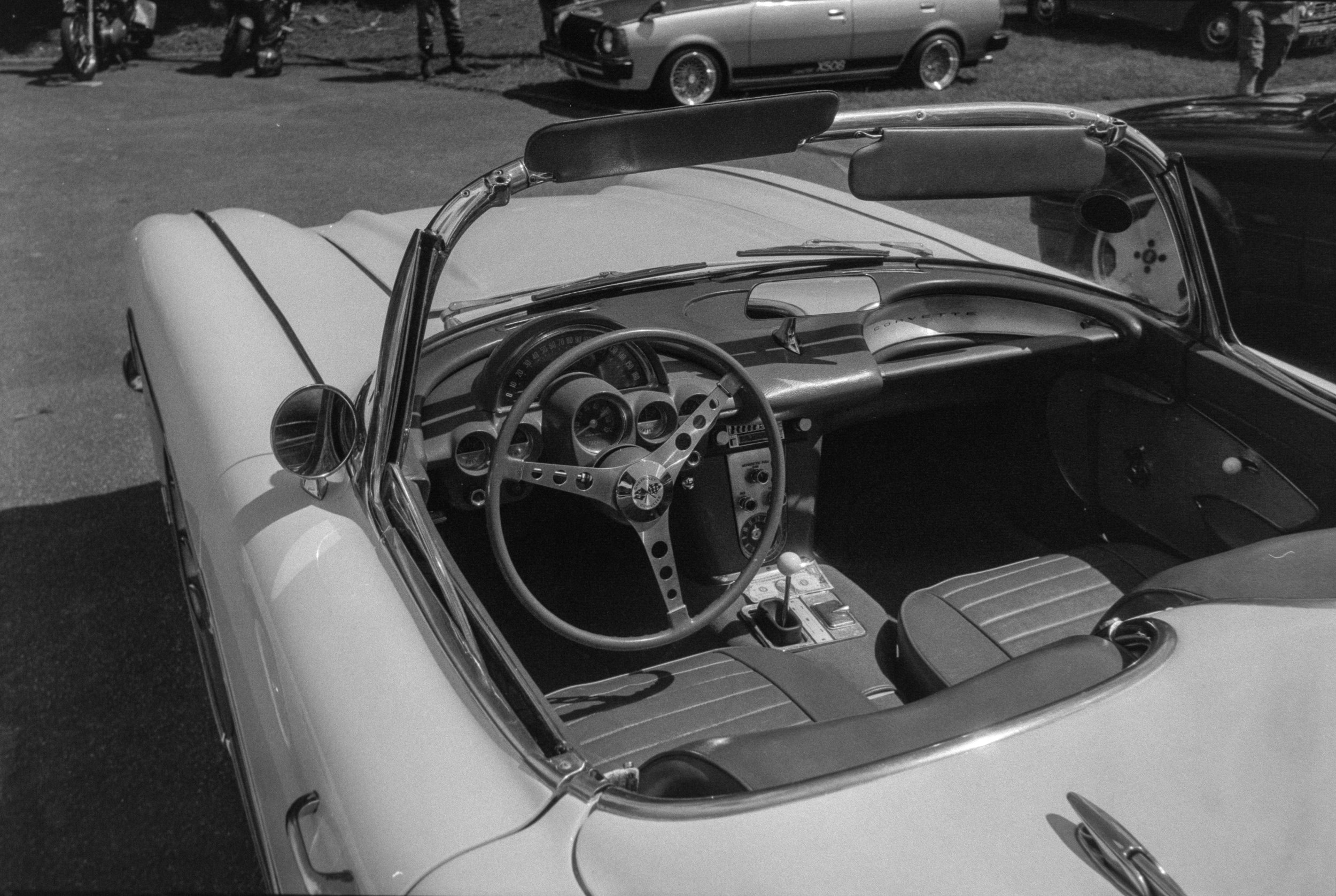 A black and white photo of the interior of a classic car with a striped dashboard and round mirrors. Other vintage cars and motorcycles are visible in the background, parked in a lot.