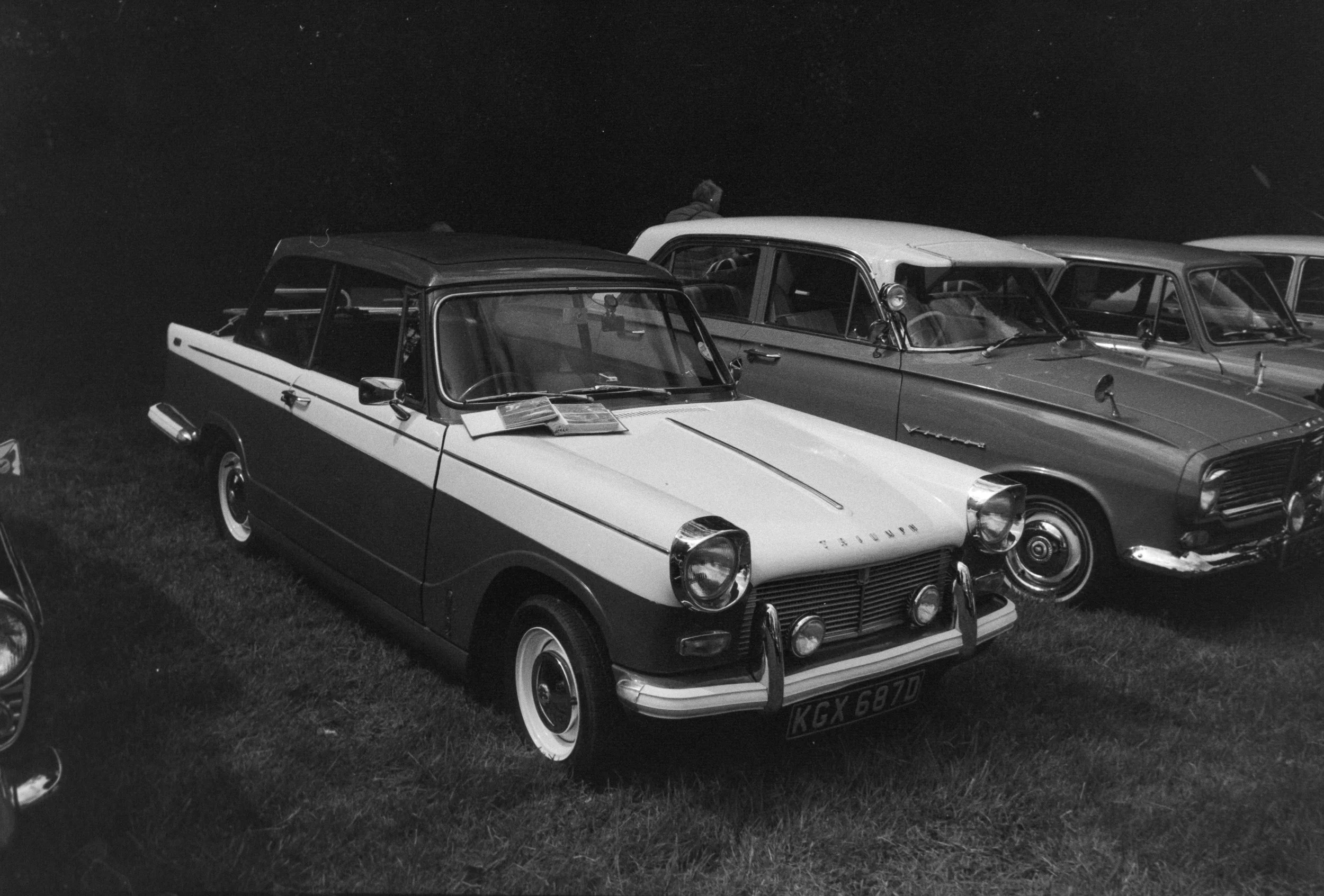Black and white photo of vintage cars parked on grass during night, with a person standing near one car.
