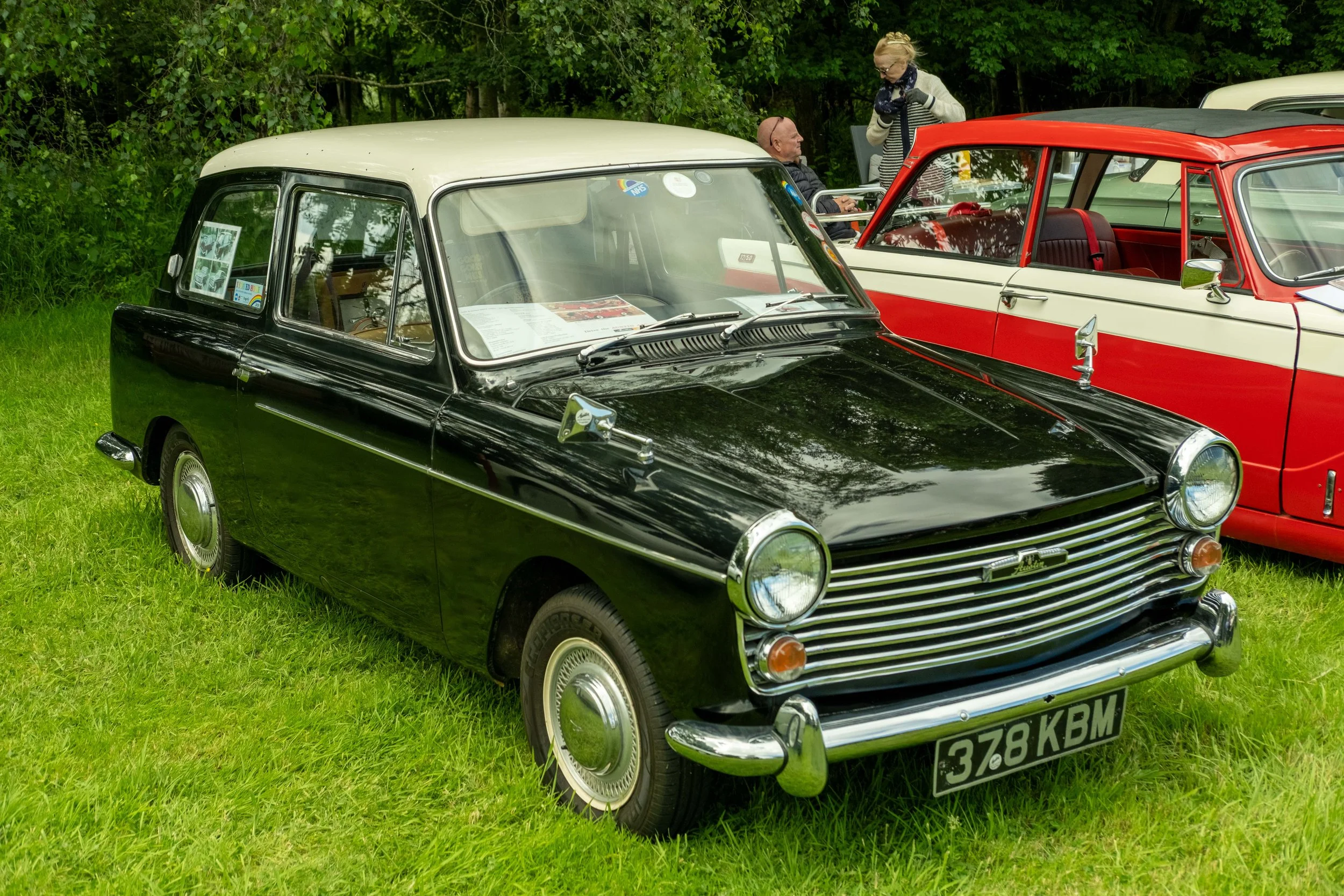 A black vintage Mini car with a white roof parked on grass at a car show, with people in the background, including a man in a wheelchair and a woman standing next to him.