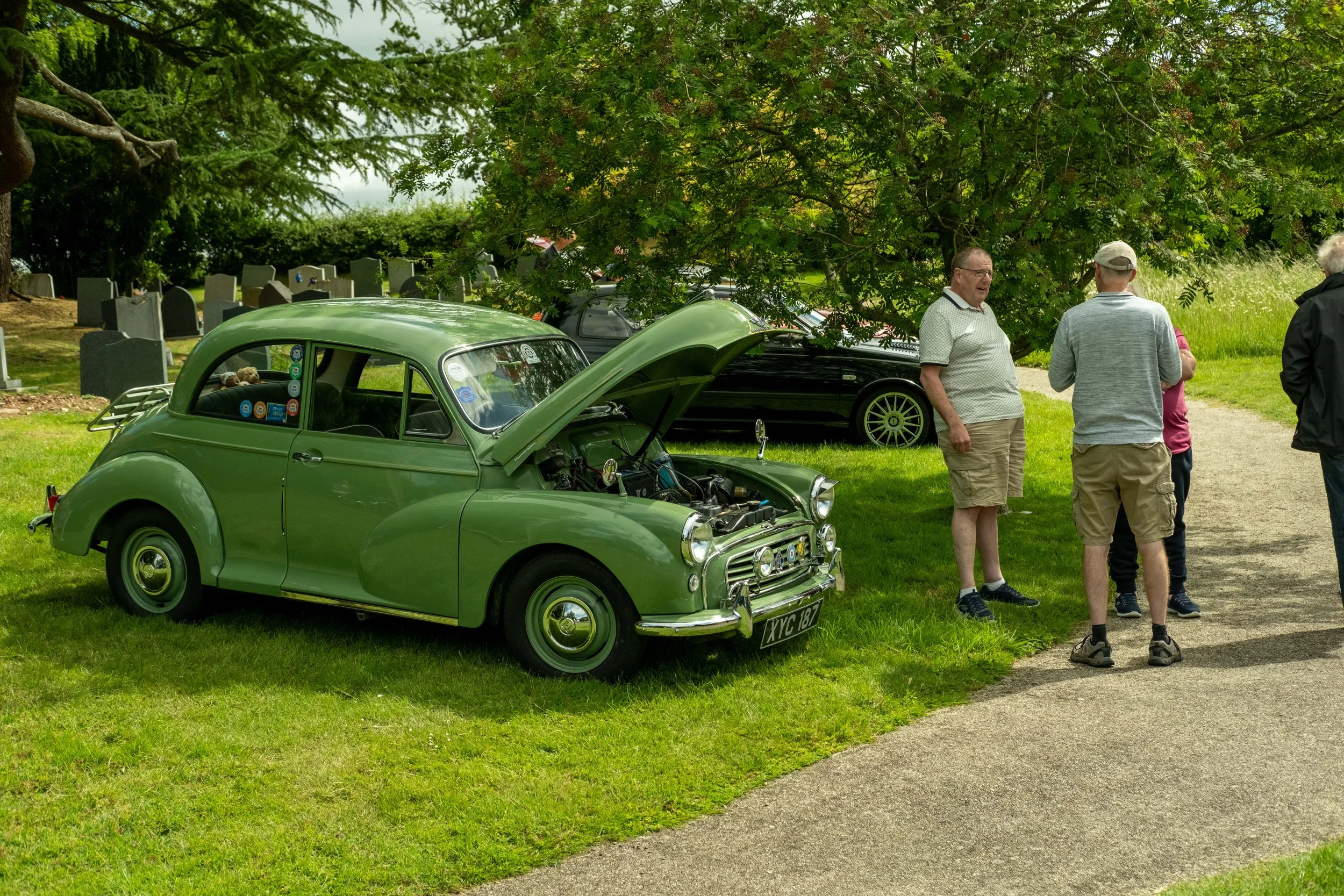 A vintage green car with its hood open parked on grass near a group of people standing and talking under a large leafy tree in a park with tombstones in the background.
