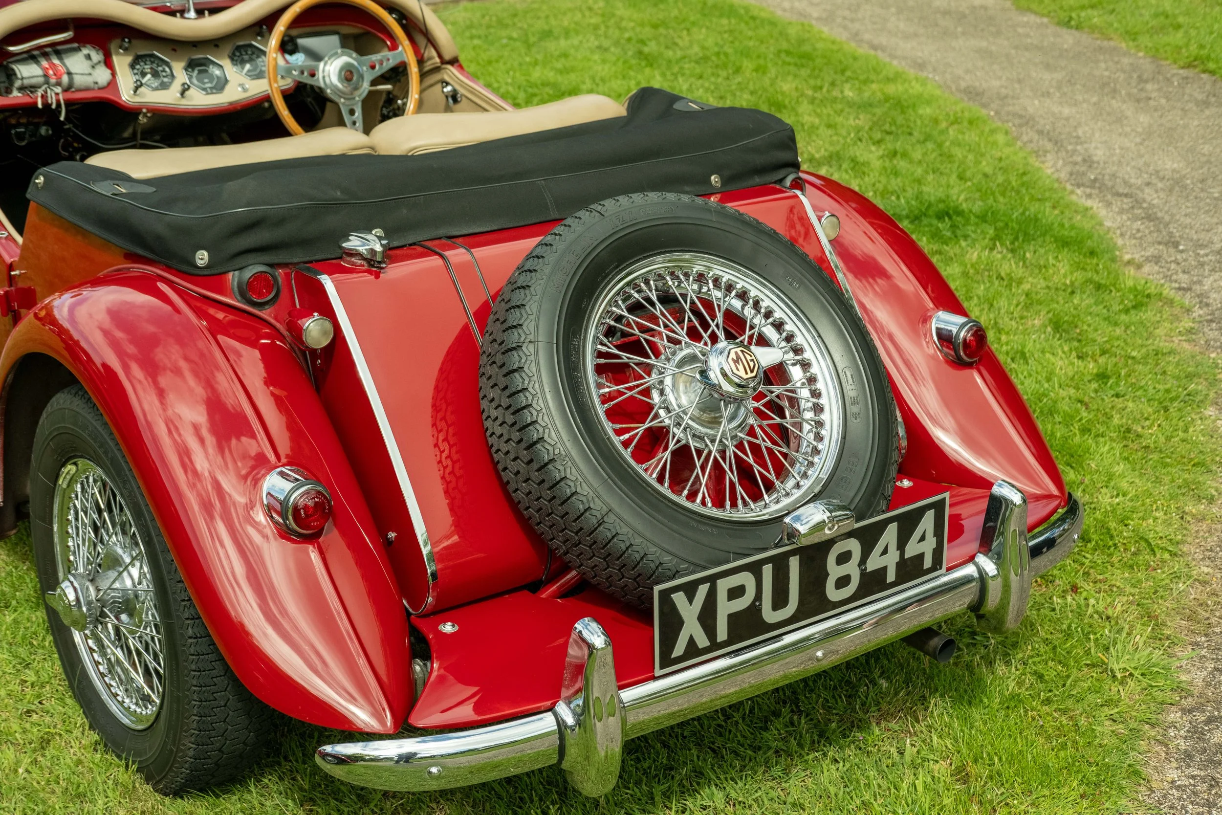 Red vintage MG sports car with a spare tire mounted on the back, parked on grass near a gravel path.