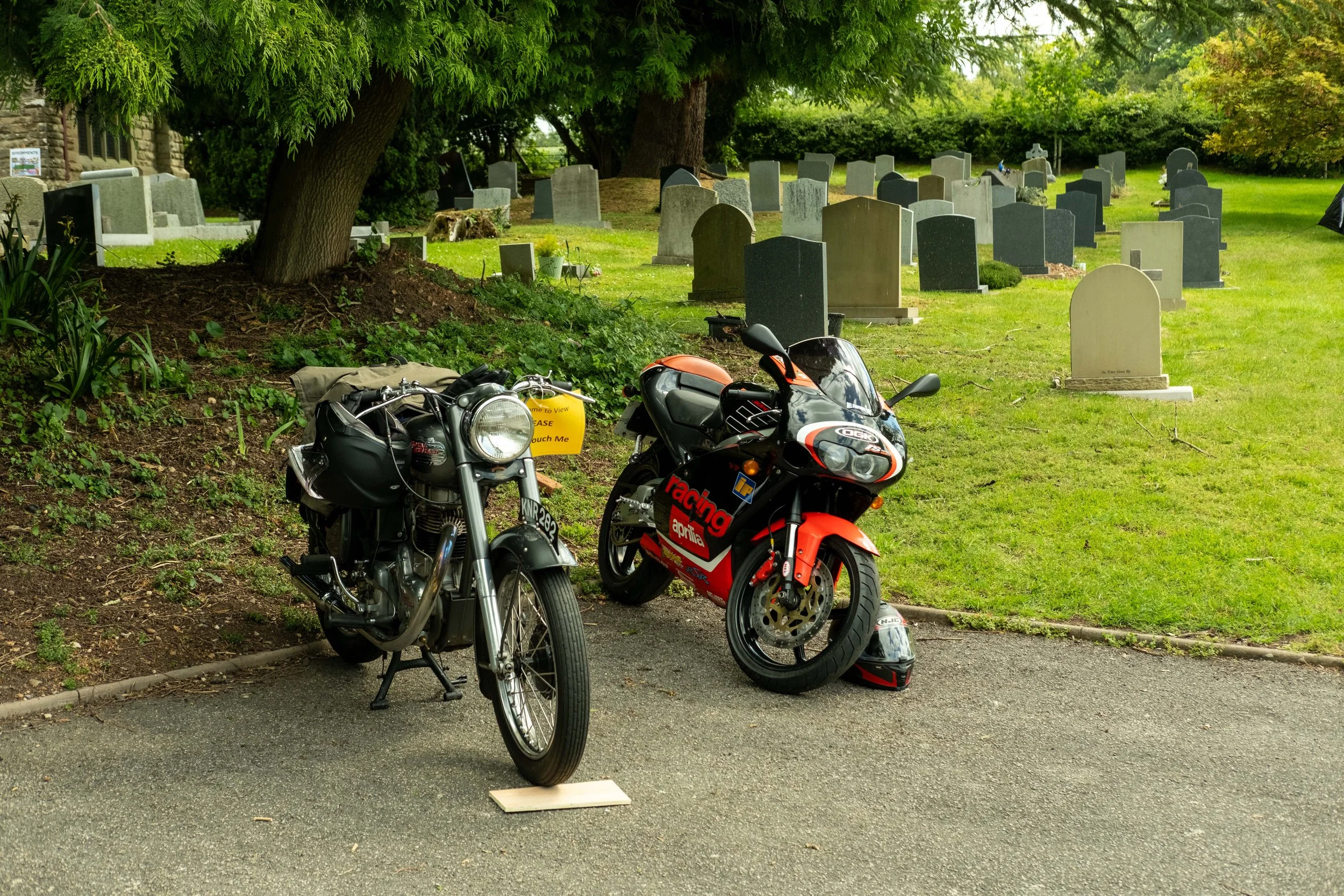 Two black and red motorcycles parked on the side of a road near a cemetery with headstones, trees, and greenery in the background.