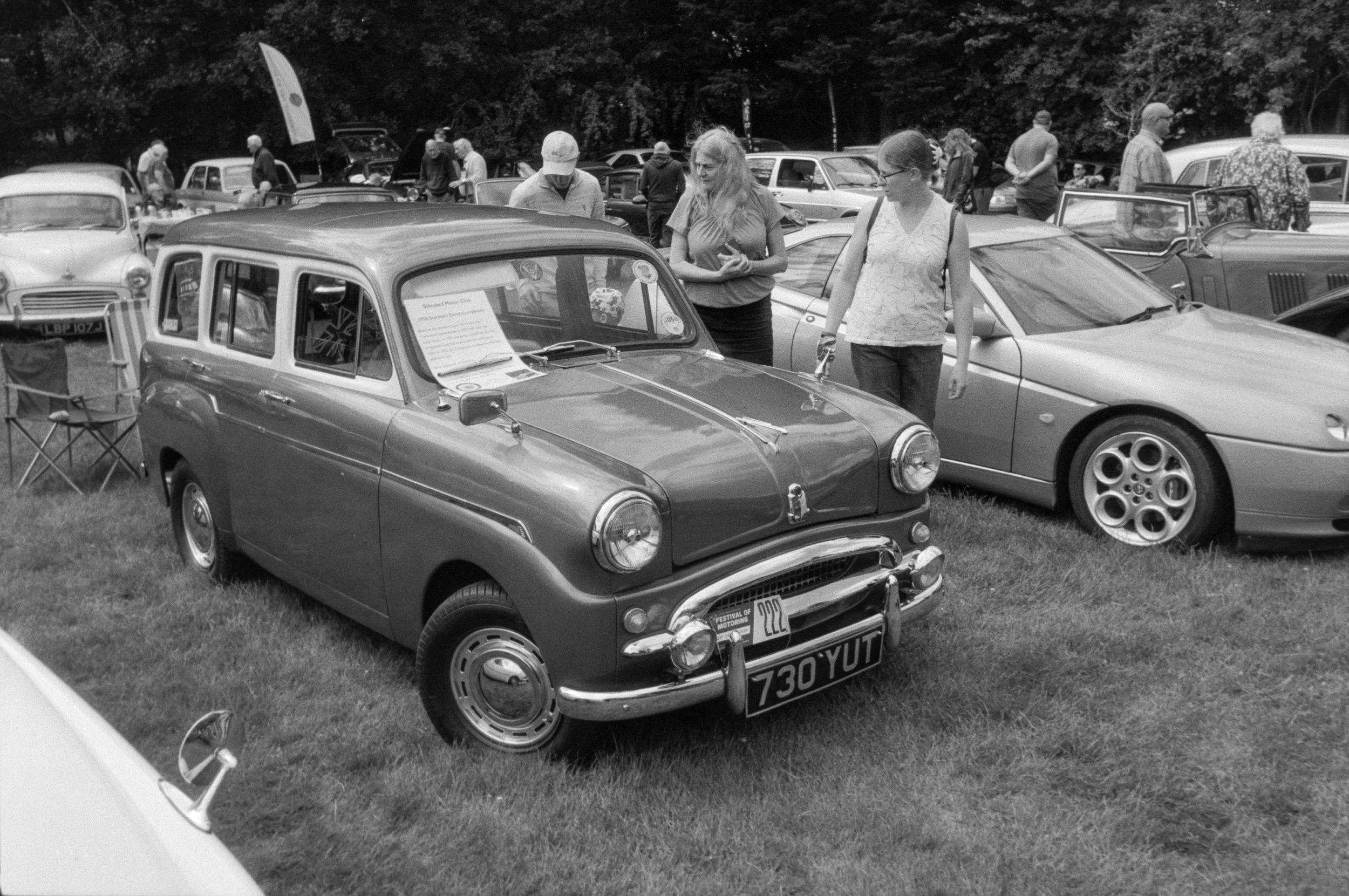 Black and white photo of a vintage car show with many classic cars and people walking and inspecting the vehicles.