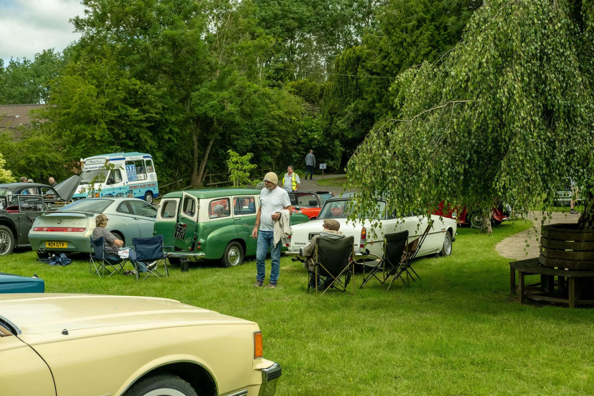 People gather around vintage cars in a park-like setting with lush green trees. Some are seated on folding chairs, others stand, and there is a food truck in the background. The scene appears to be a car show or outdoor event.
