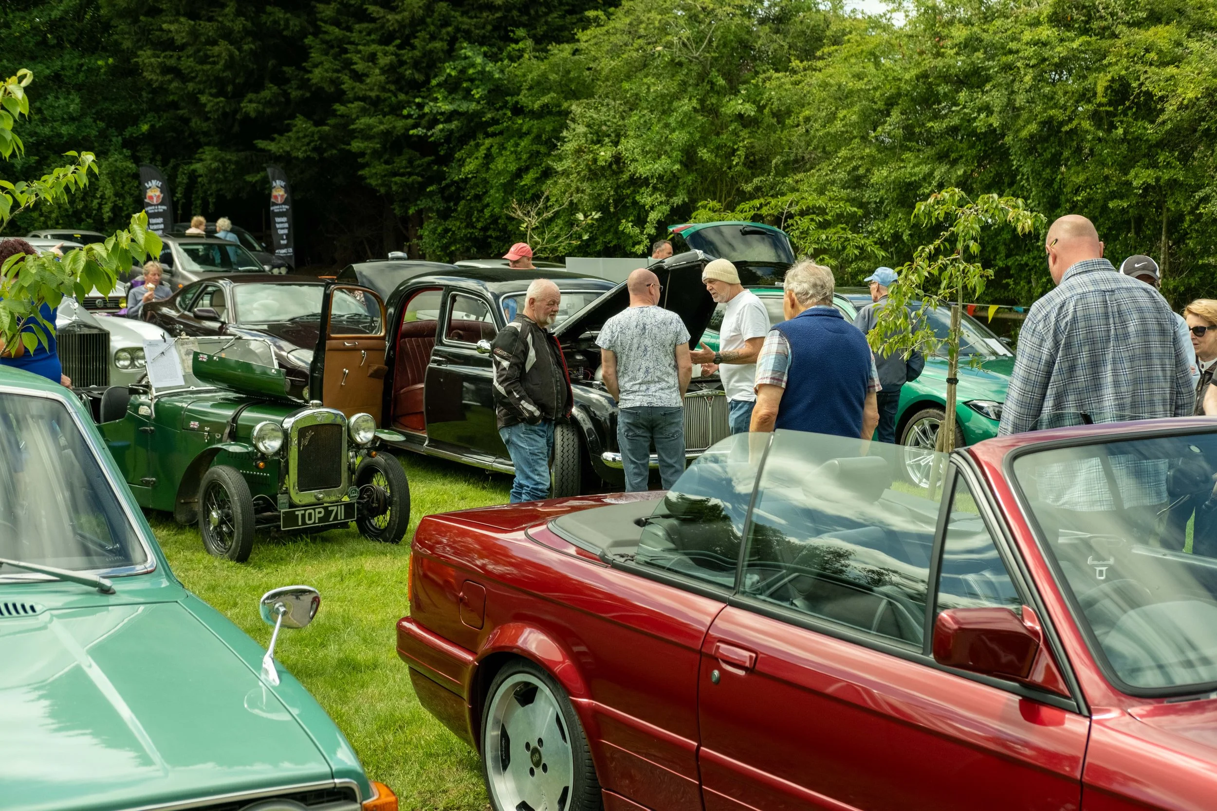 People gathered around vintage cars at an outdoor classic car show, with lush green trees in the background.