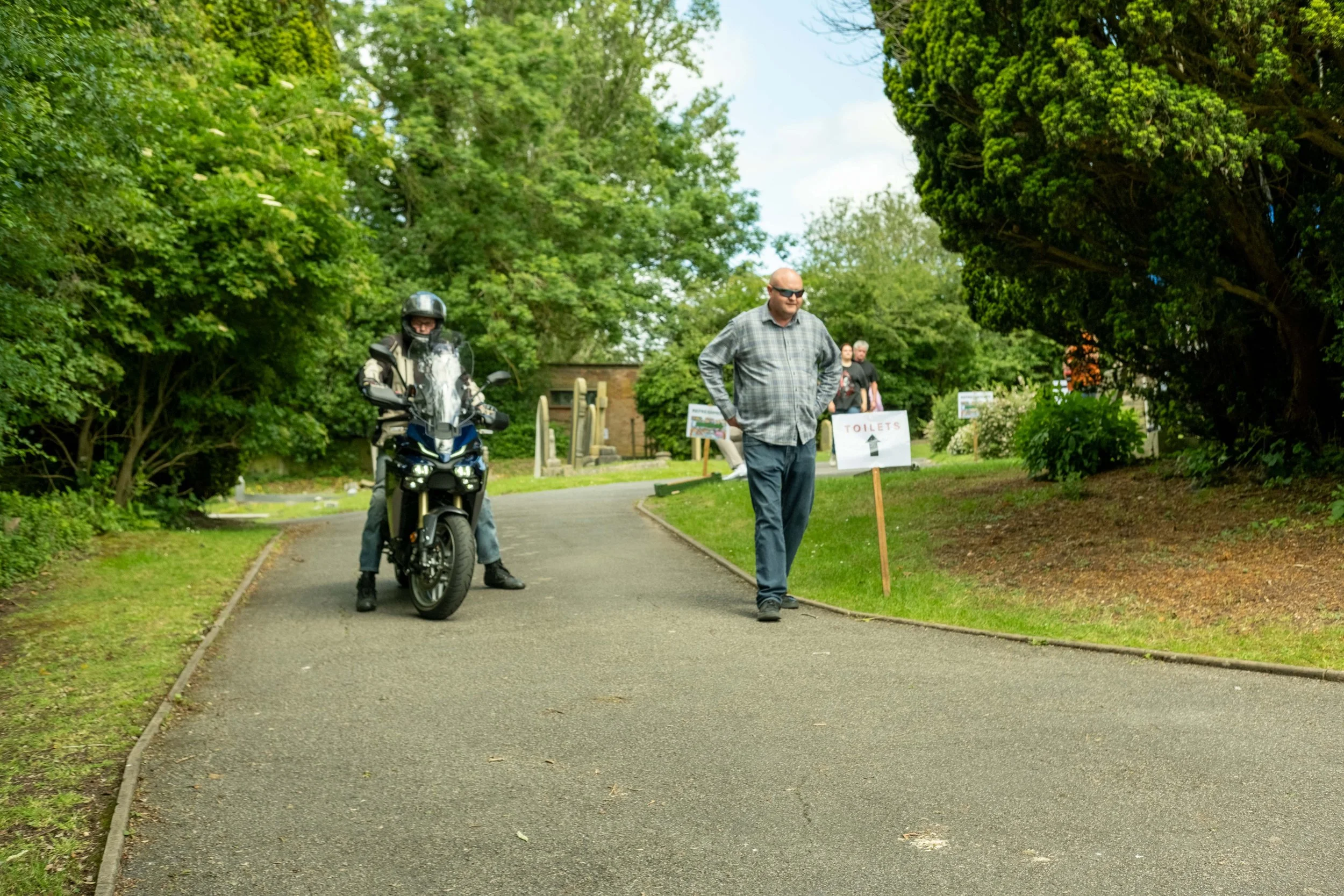 A man on a motorcycle with a helmet and a man with sunglasses walking nearby, sign pointing to toilets, in a green park with trees and a pathway