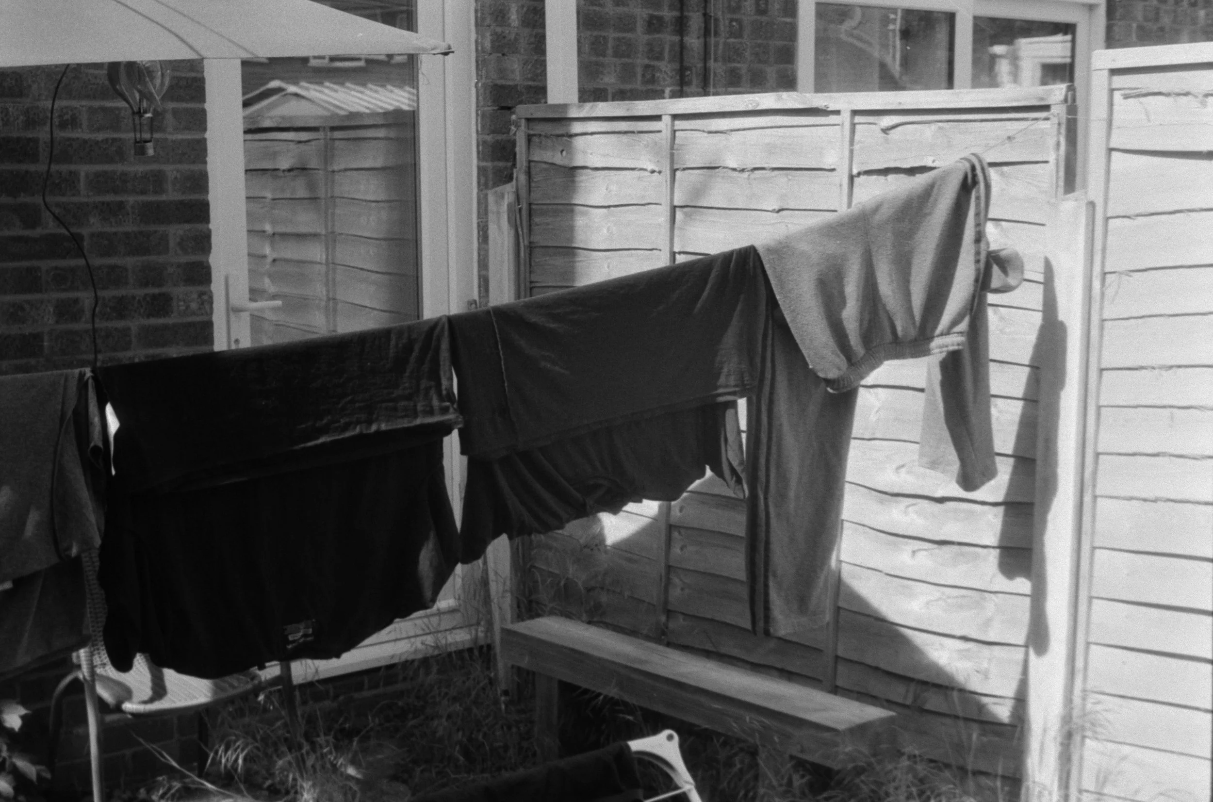Clothes hanging on a clothesline outside a house with a brick wall, window, and wooden fence, in black and white.