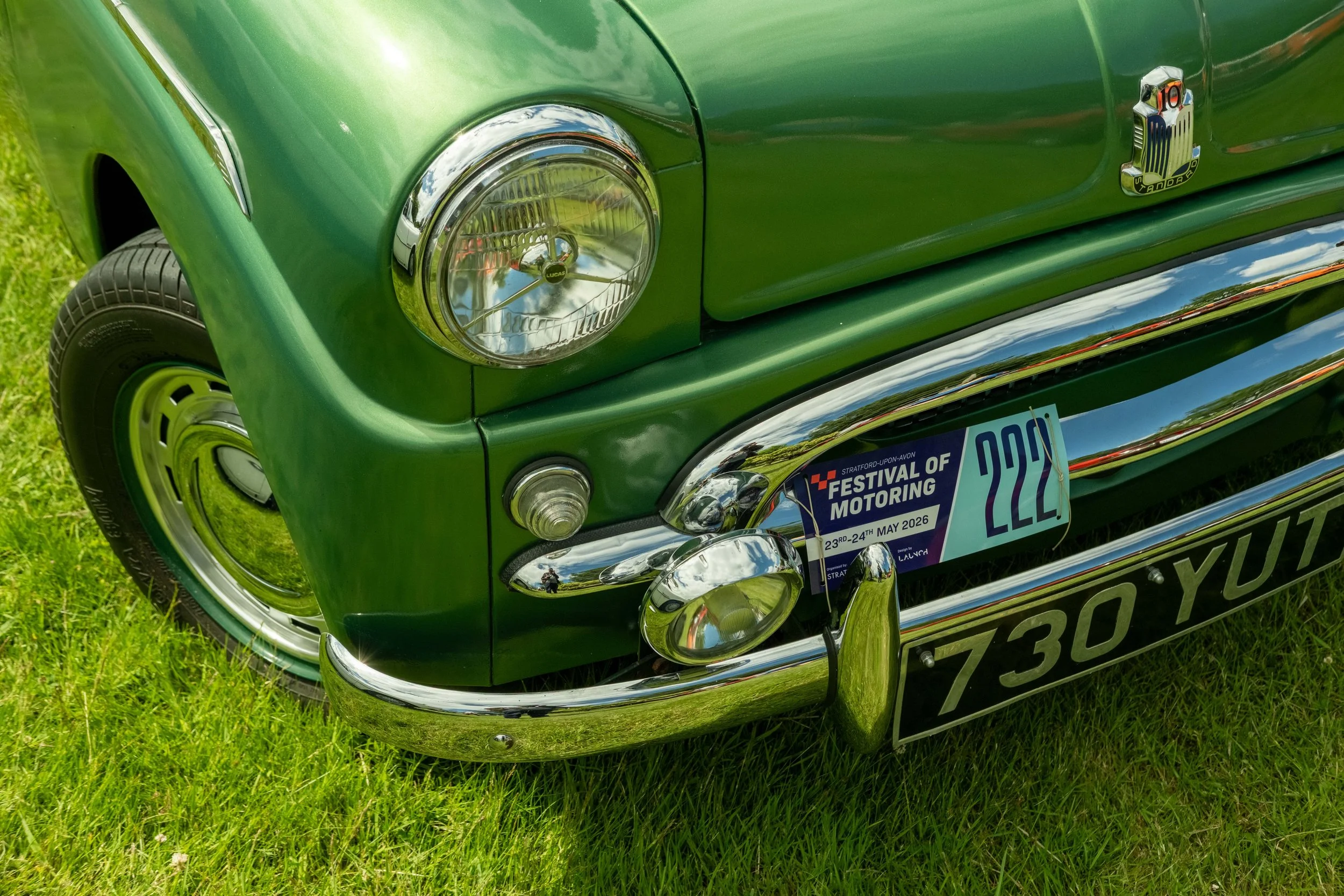 Close-up of a vintage green car's front, showing headlight, bumper, and part of a bumper sticker reading 'Festival of Motoring 23rd-24th May 2026.'