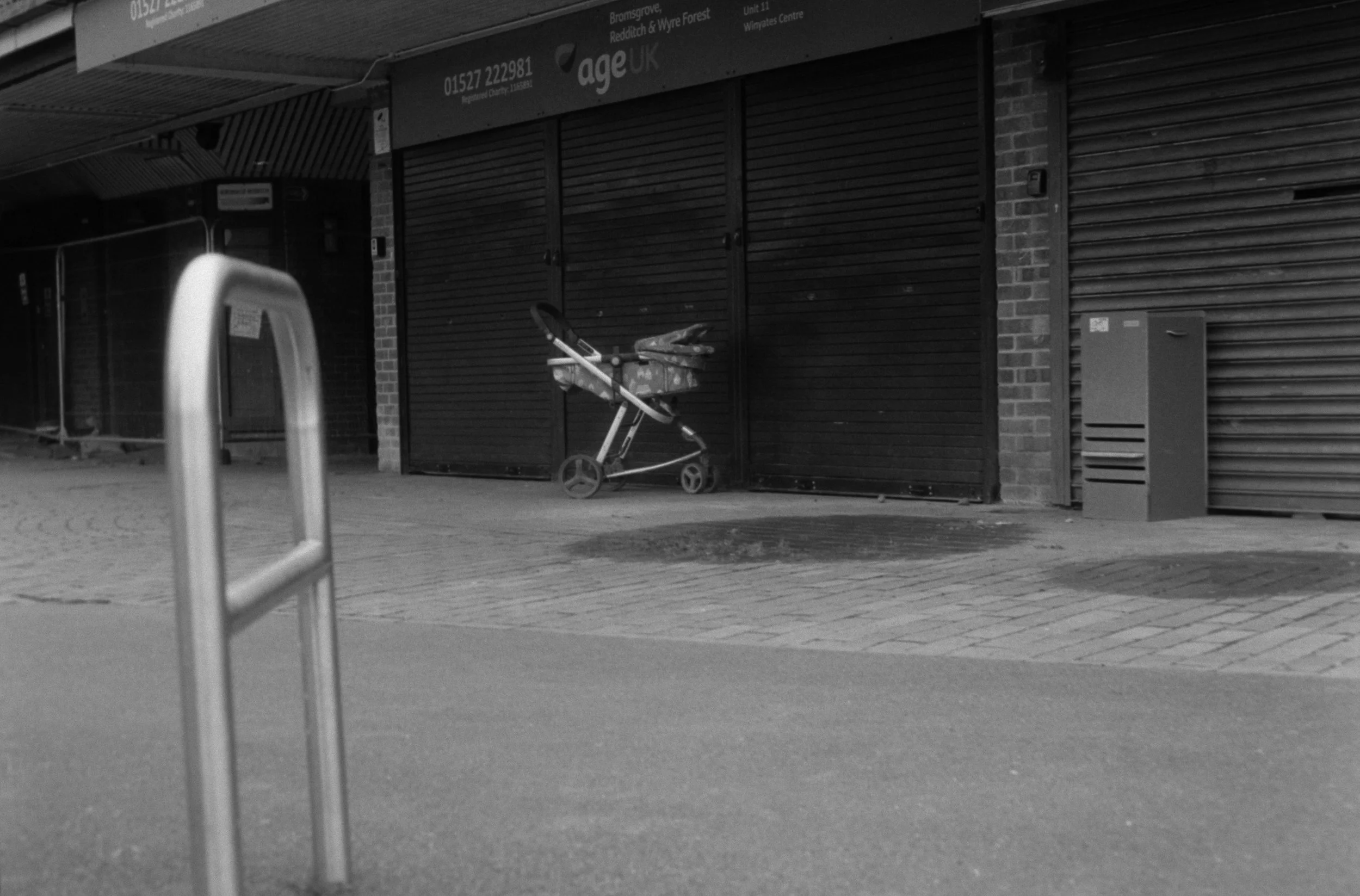 A street scene with a closed shopfront featuring black metal shutters, a shopping trolley with a bag inside, and a metal barrier in the foreground.