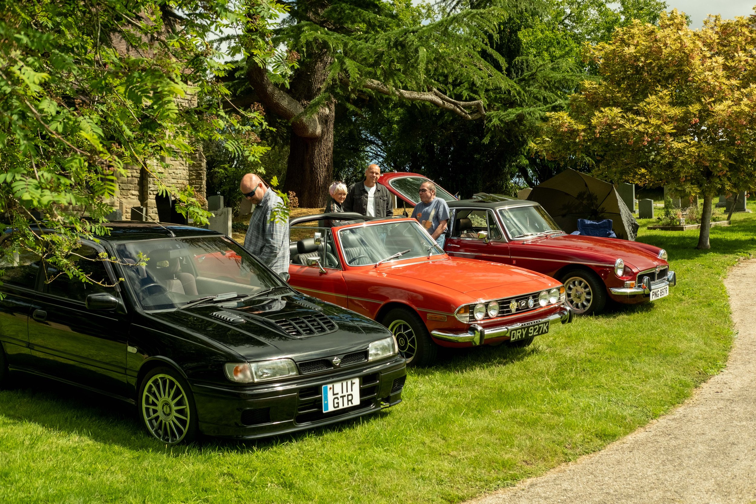 Several vintage and modern cars parked on a grassy area under trees, with people standing and conversing nearby.