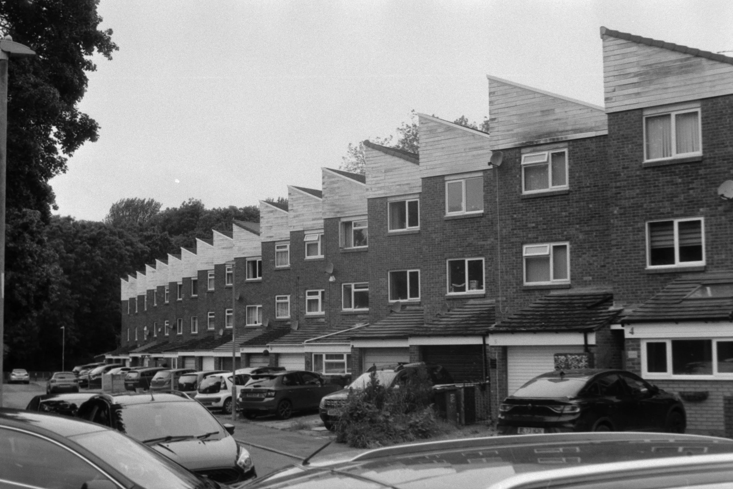 Black and white photo of multiple modern apartment buildings with triangular roof sections, parked cars in front, and trees in the background.