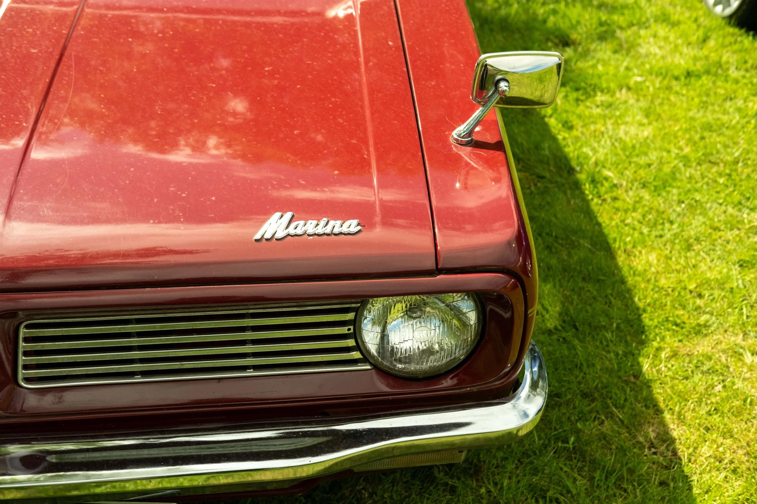 Close-up of the front of a vintage red car with a chrome side mirror and a 'Mauna' emblem, parked on grass