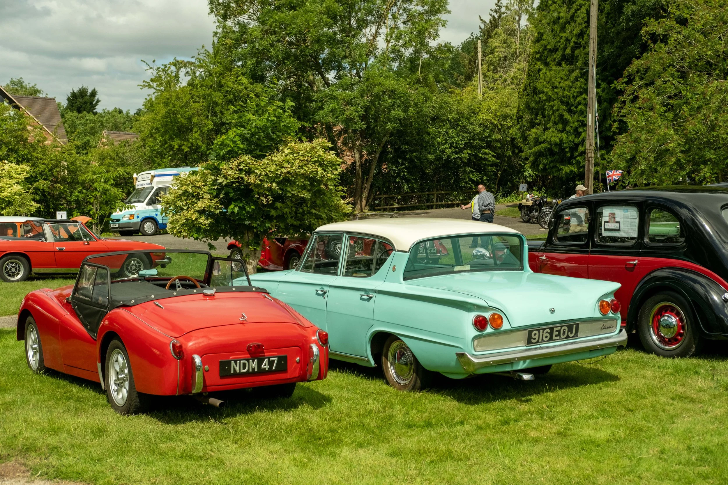 Various vintage cars parked on a grassy area with trees and electric poles in the background. Some cars have Union Jack flags, and people are walking nearby.