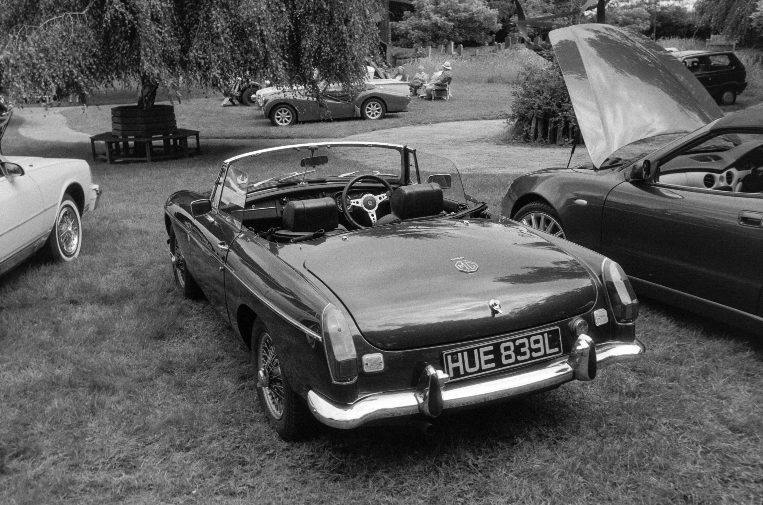 Black and white photo of a vintage convertible sports car with the hood open, parked on a grassy field. Other parked cars and people sitting on chairs are in the background.