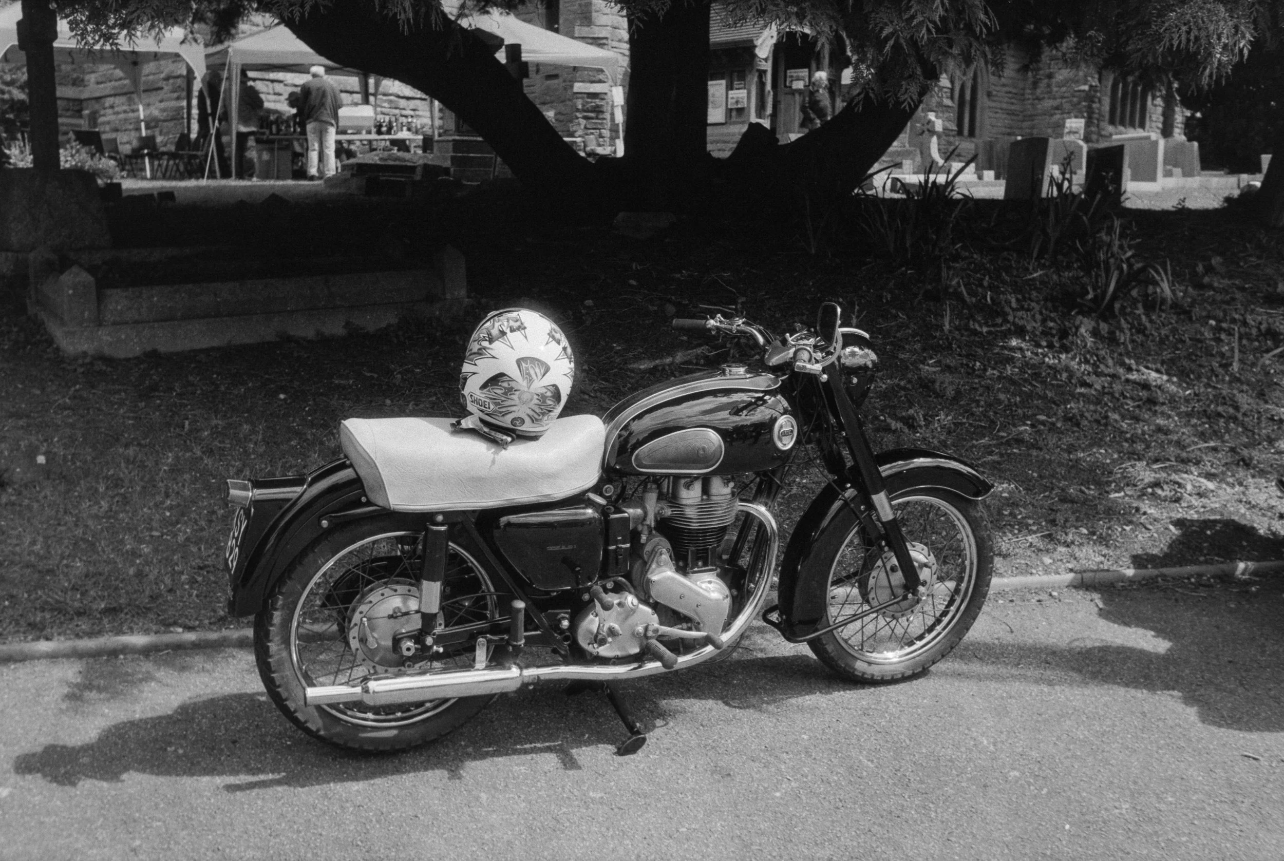 A vintage motorcycle parked on the side of a street, with a helmet on the seat. In the background, there is a park with a large tree, benches, and a building with a couple of people near outdoor tables.